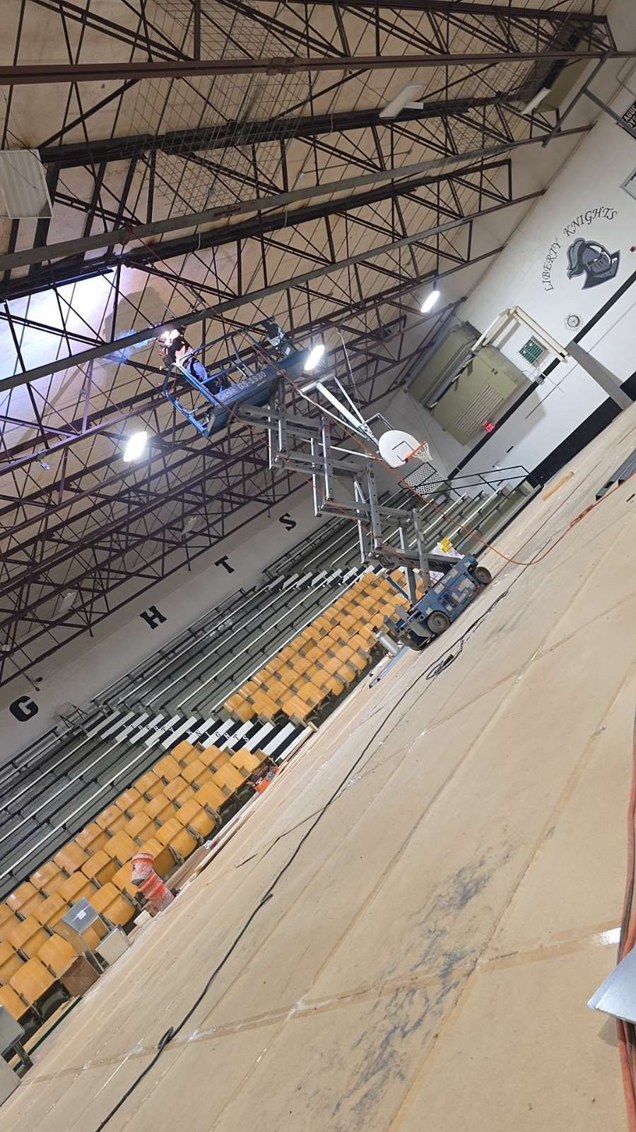 Indoor gymnasium with bleachers, basketball hoop, and high ceiling lights, viewed at an angle.