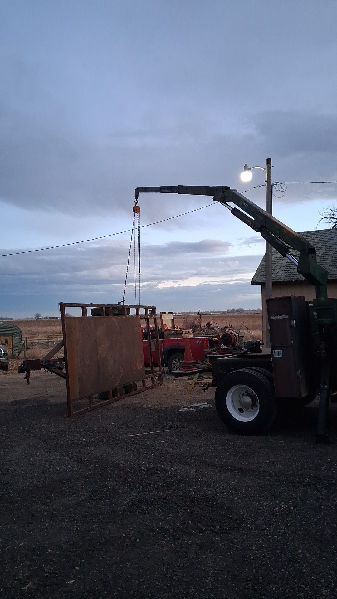 Farmyard at dusk with a tractor-mounted crane lifting a hanging load near string lights and a wooden pen.