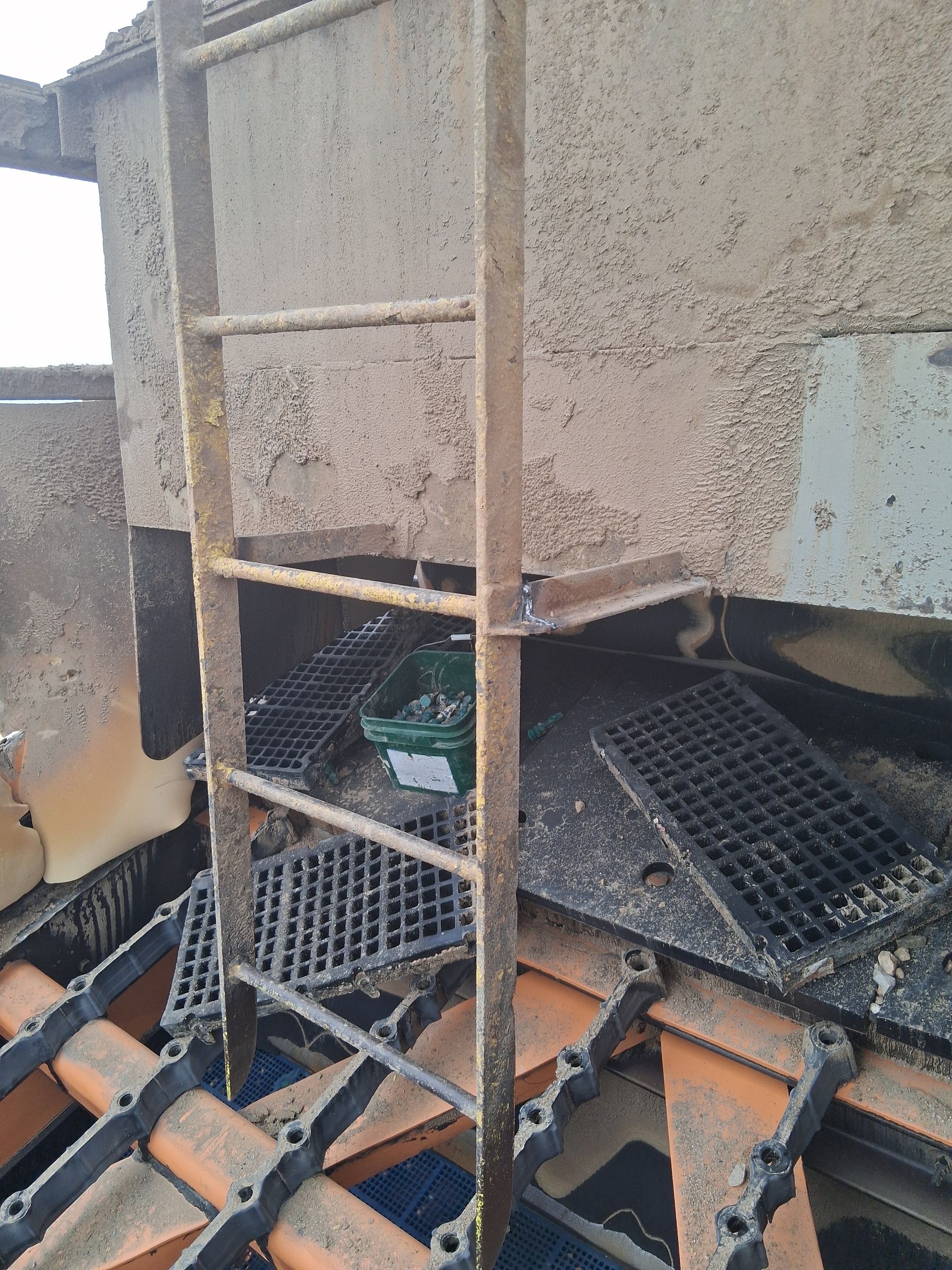 Rusty metal ladder beside industrial machinery and grates, with a green crate below.