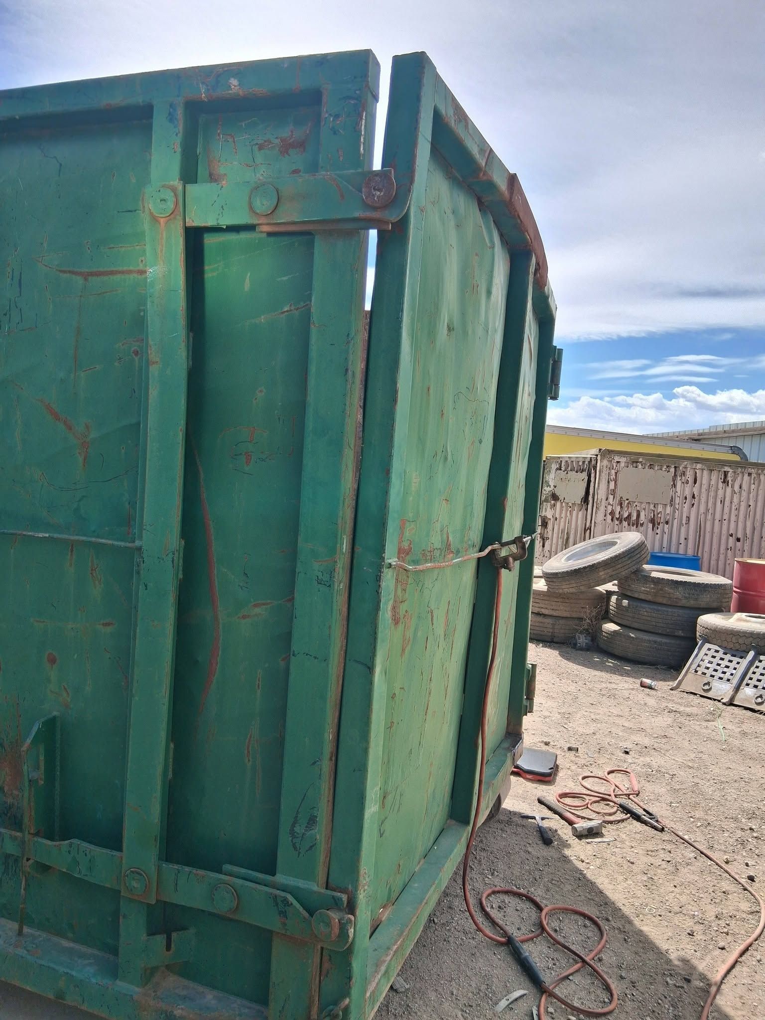 Green metal dumpster on a gravel lot with a fence, hose, and scattered tools nearby
