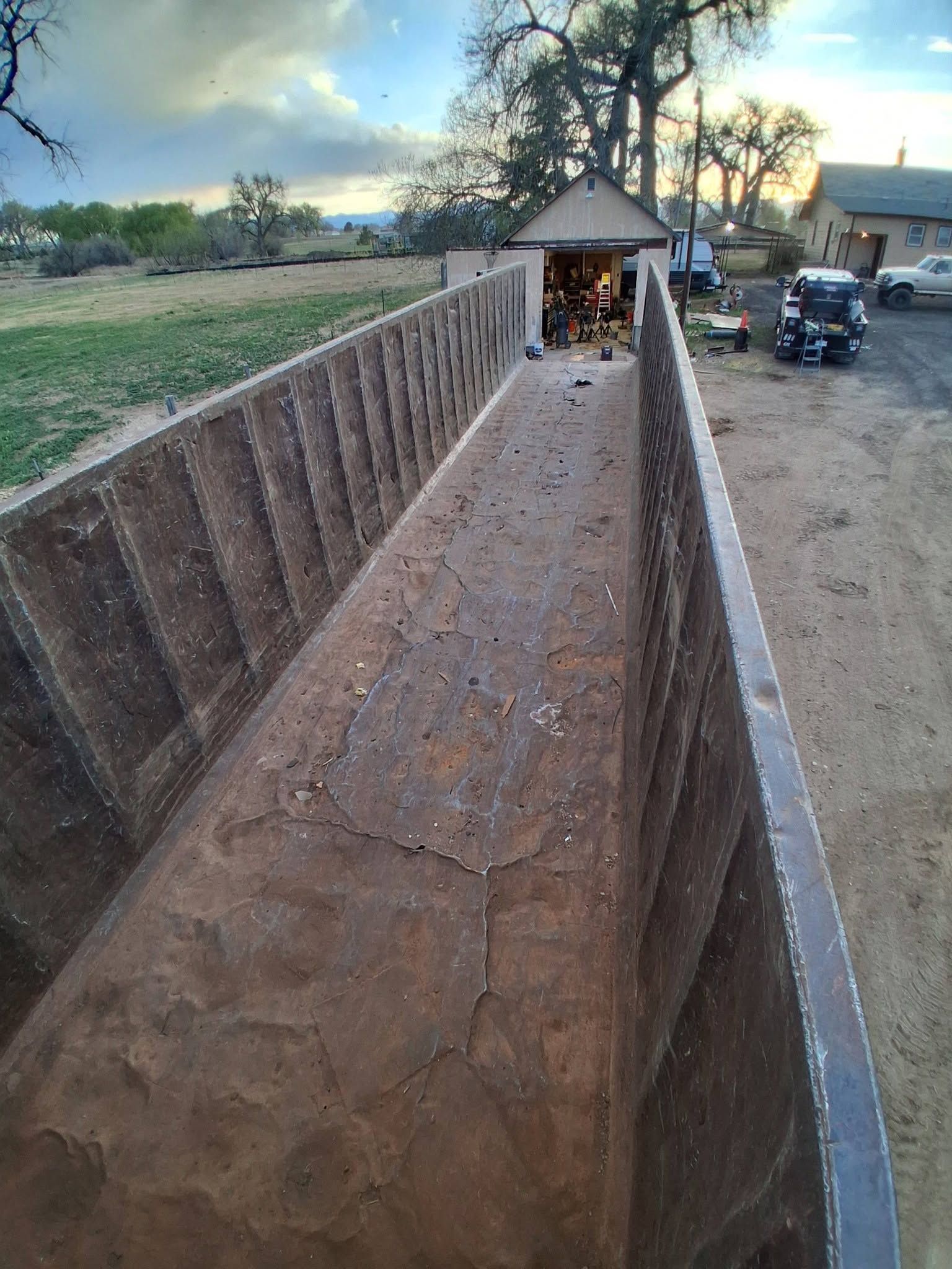 Long rusted metal trough or chute leading to a small shelter in a rural yard at dusk