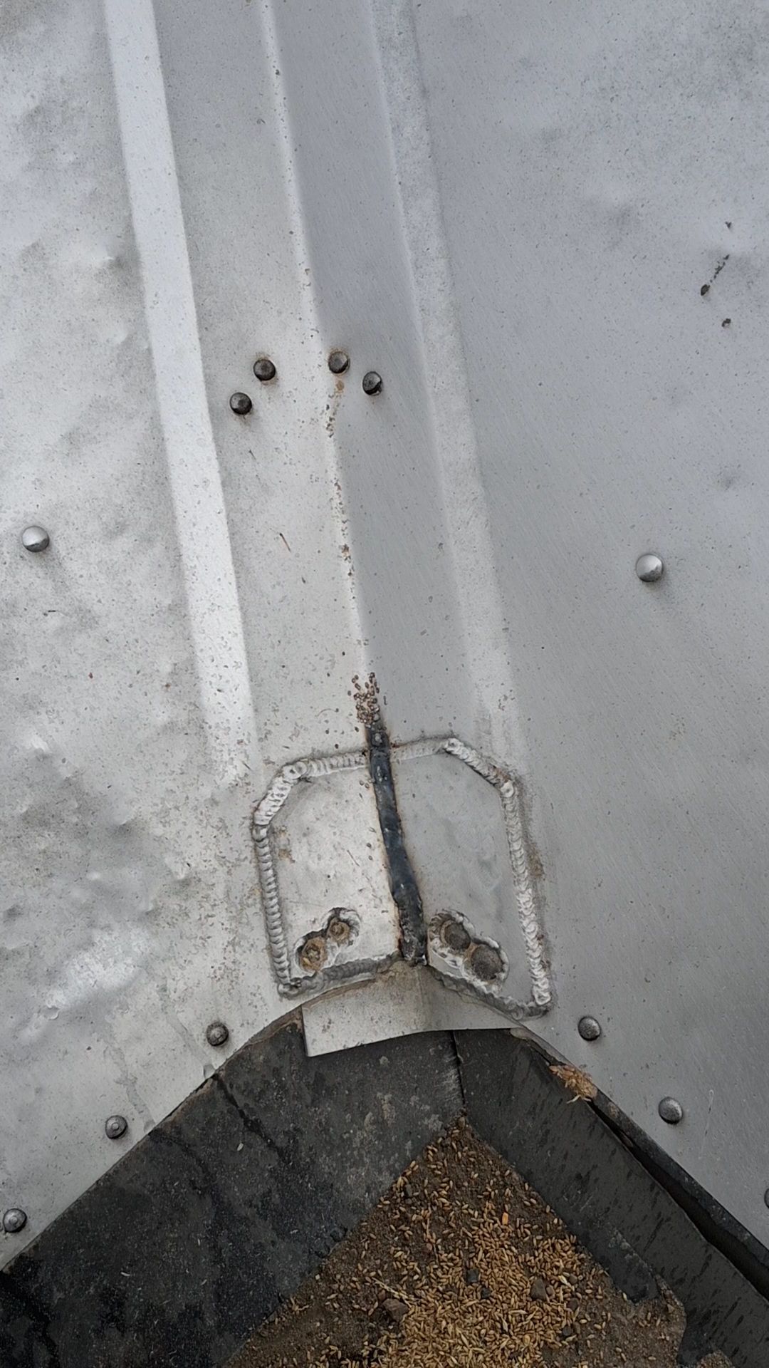 Close-up of a gray metal surface with rivets and a corroded seam at the bottom corner.