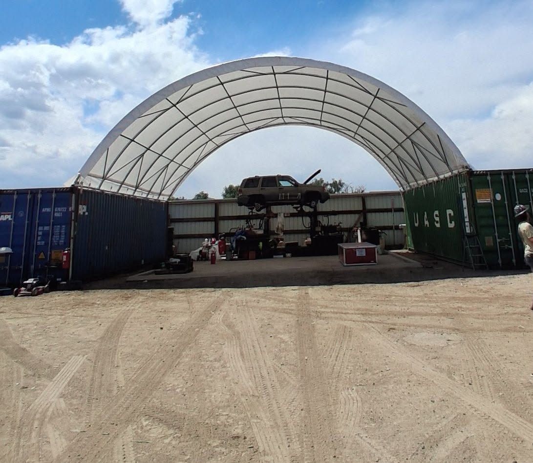 Large white arched tent in a dirt yard, with shipping containers on both sides and equipment inside.