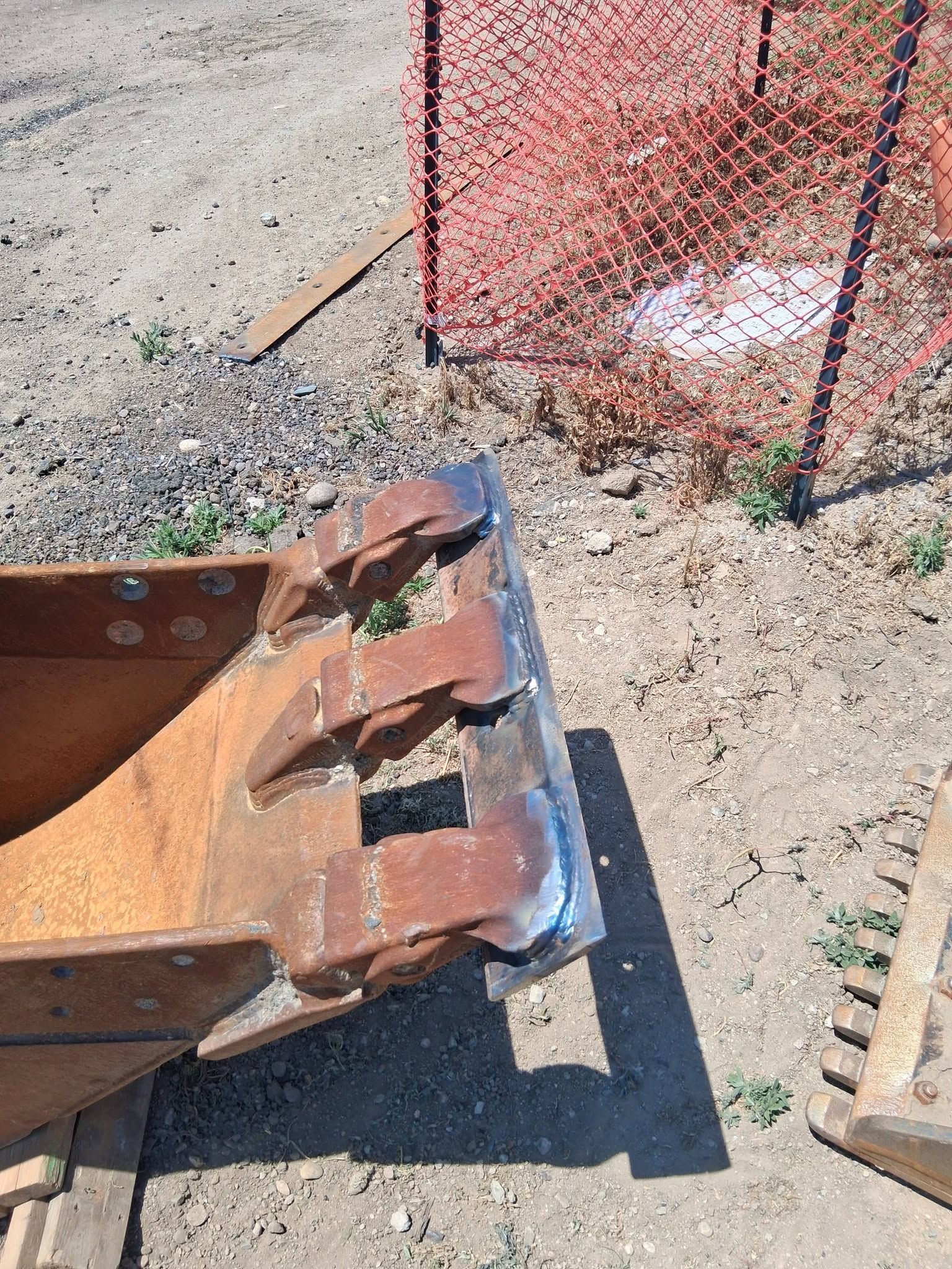 Rusty metal dump bin on gravel beside orange safety fencing and wooden planks.