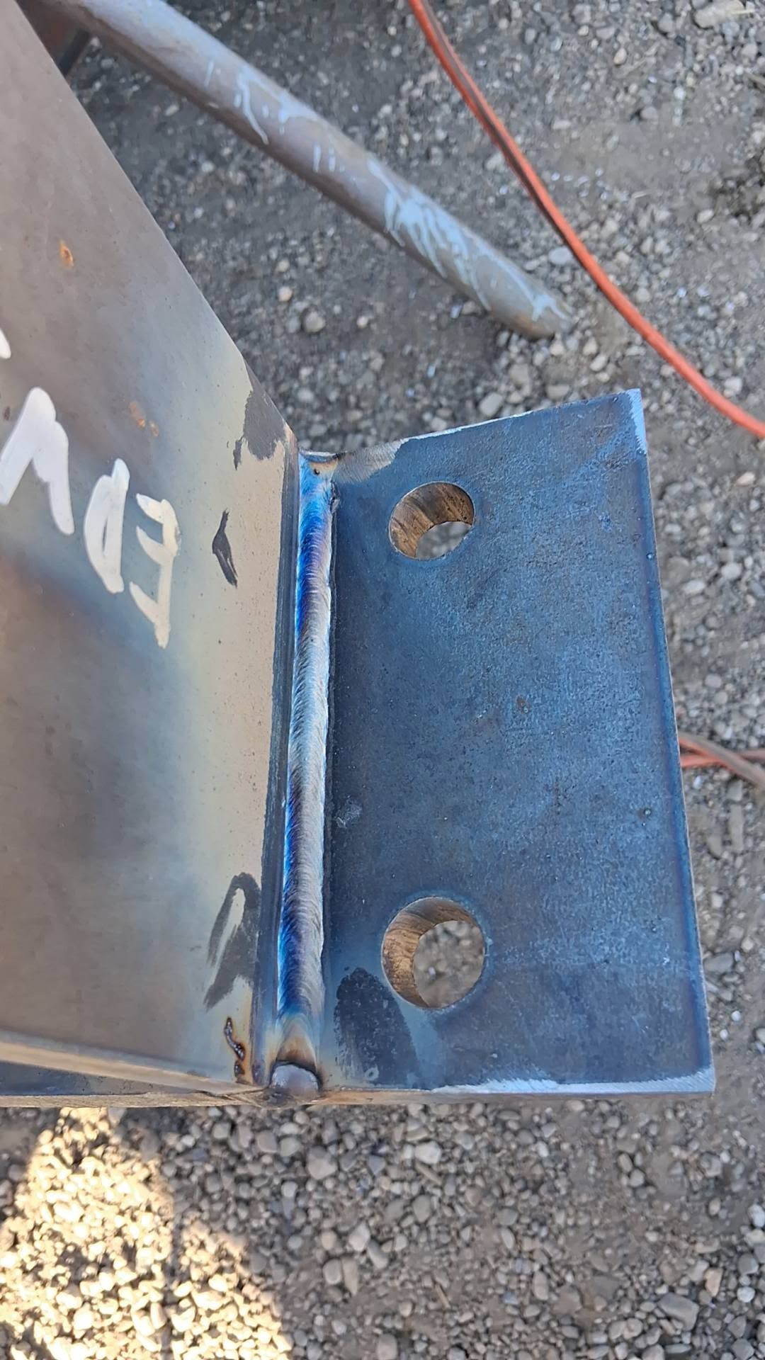 Close-up of a blue metal bracket bolted to a rusty panel on gravel ground.
