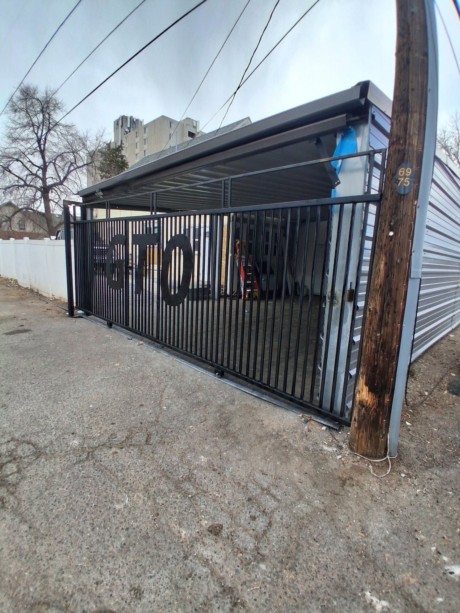 Black metal carport gate beside a gravel alley, with a wooden utility pole and building in the background
