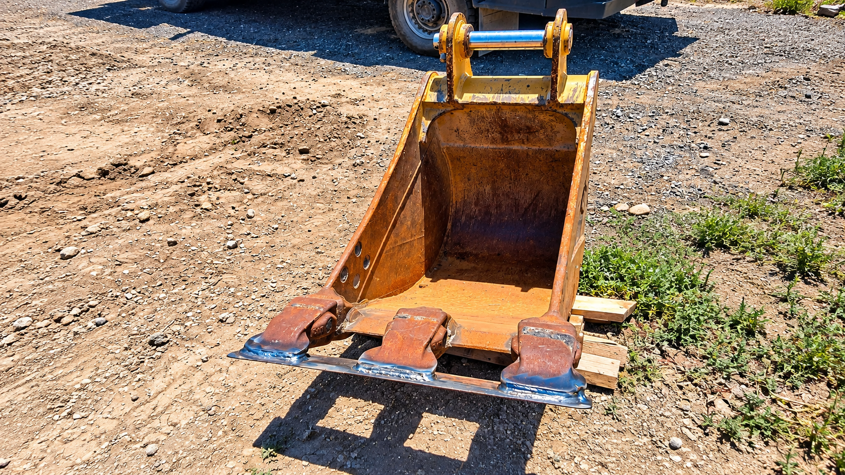 Rusty excavator bucket on gravel, with dirt inside and small wooden blocks beneath it.