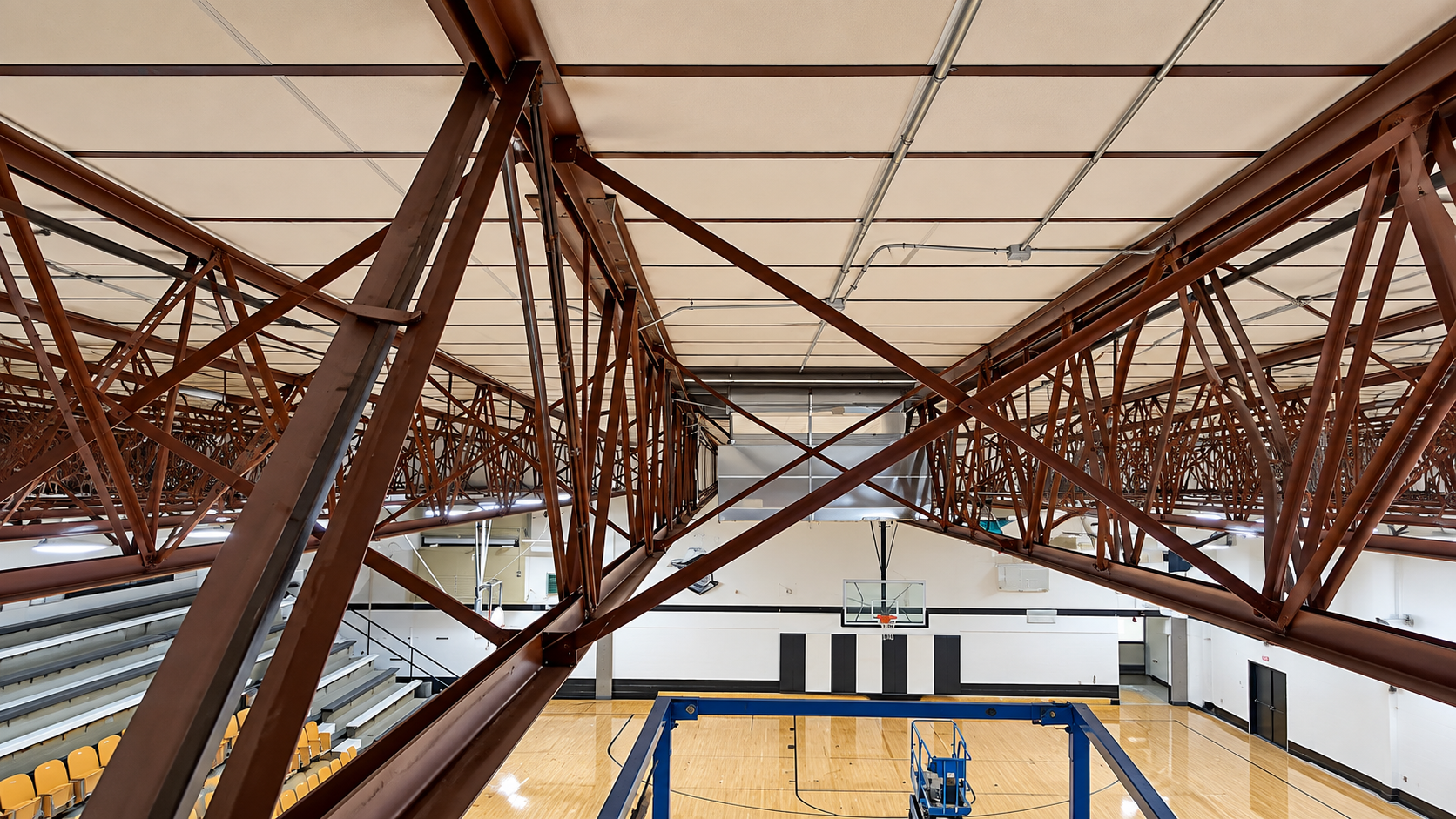 Interior view of a gymnasium roof with exposed brown steel trusses above a basketball court
