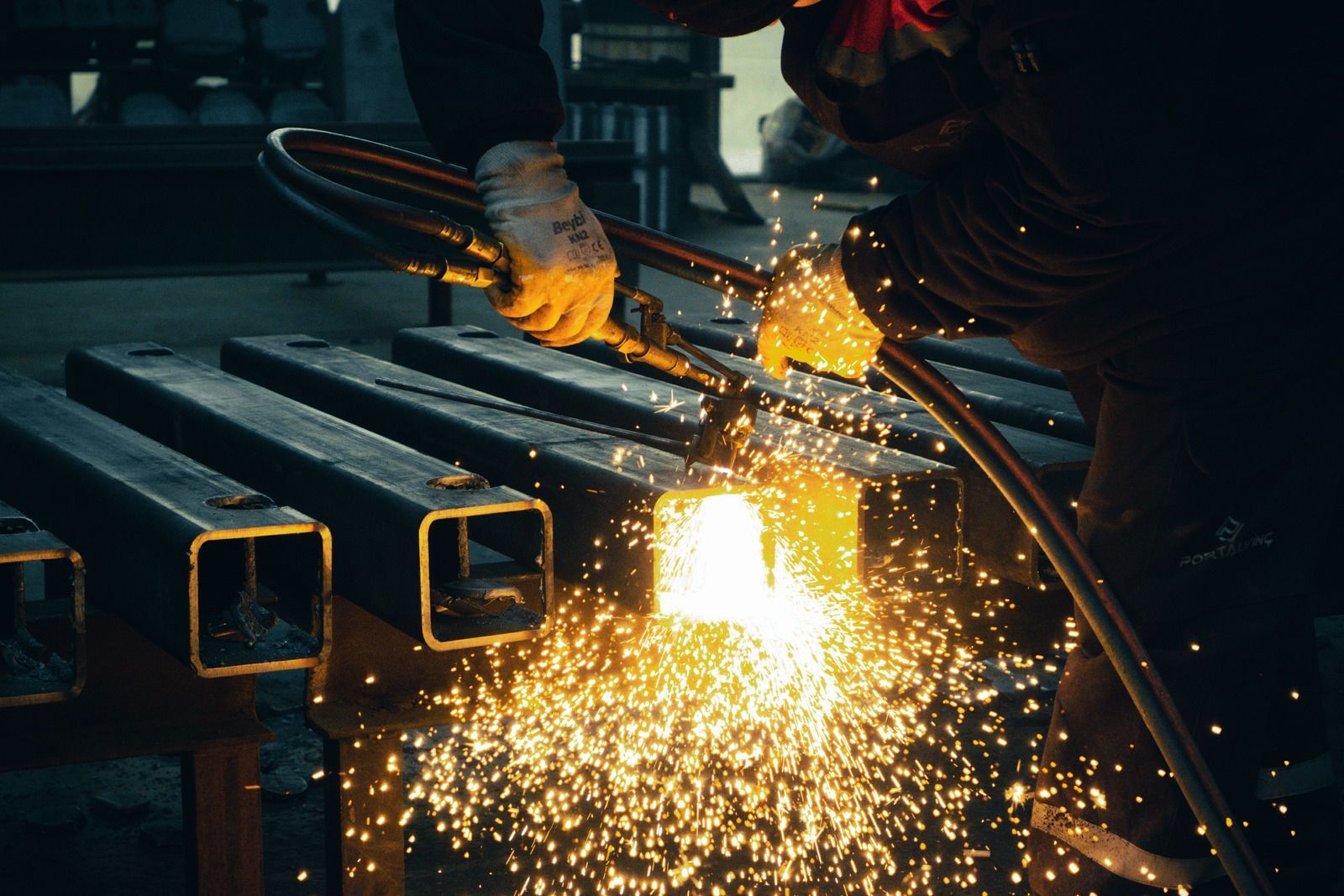Worker cutting metal pipes with a grinder, sending bright sparks across a dark workshop.