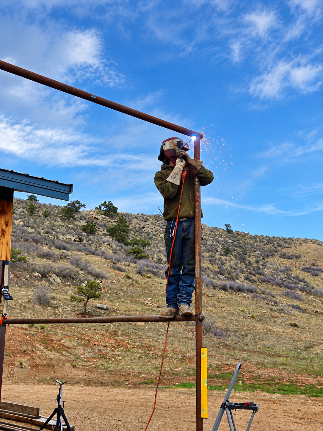 Worker standing on a metal frame outdoors, using a tool on a horizontal pipe under a blue sky