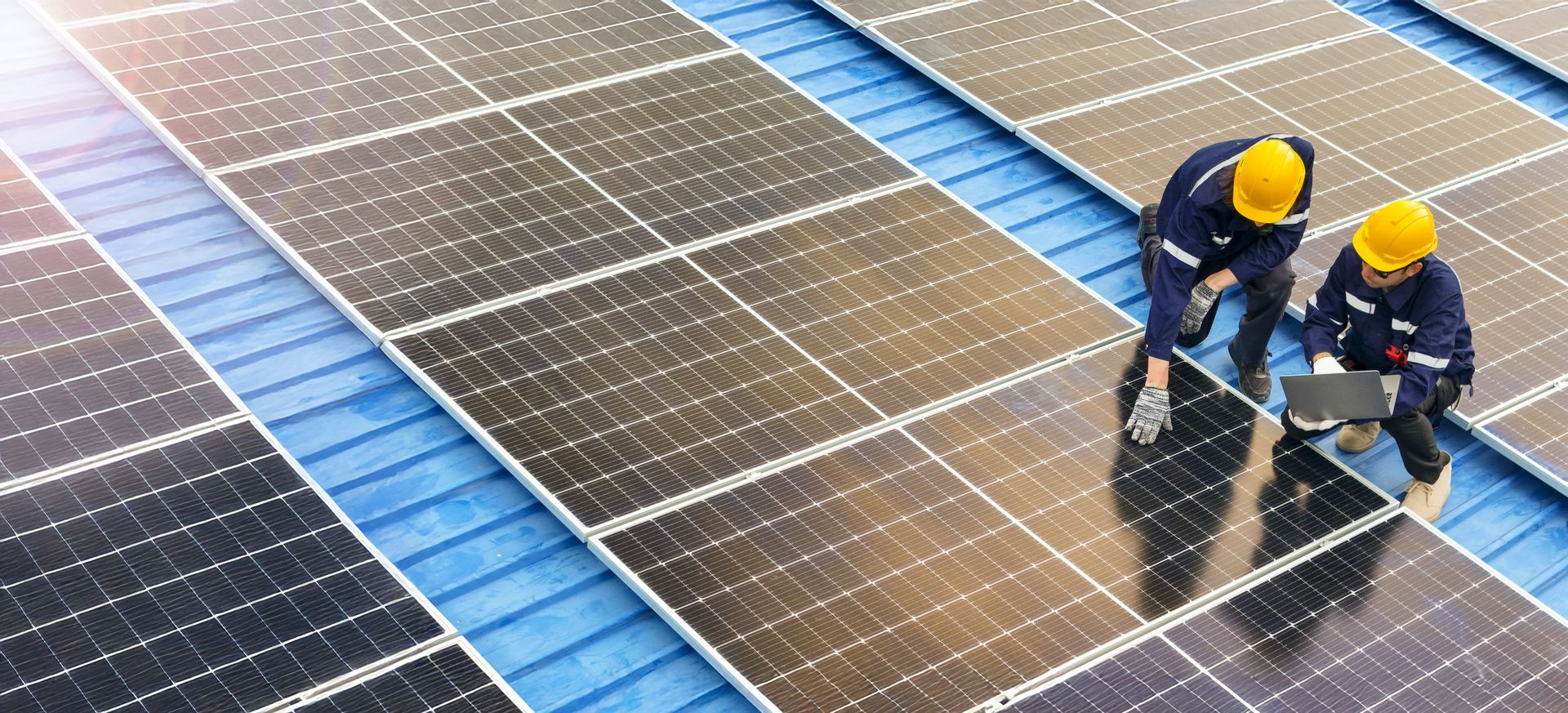 Deux techniciens, casqués et équipés de protections, inspectent des panneaux solaires installés sur un toit métallique bleu.