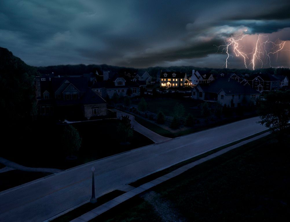 Un quartier résidentiel la nuit, sous un ciel sombre et nuageux, avec un éclair brillant au loin.