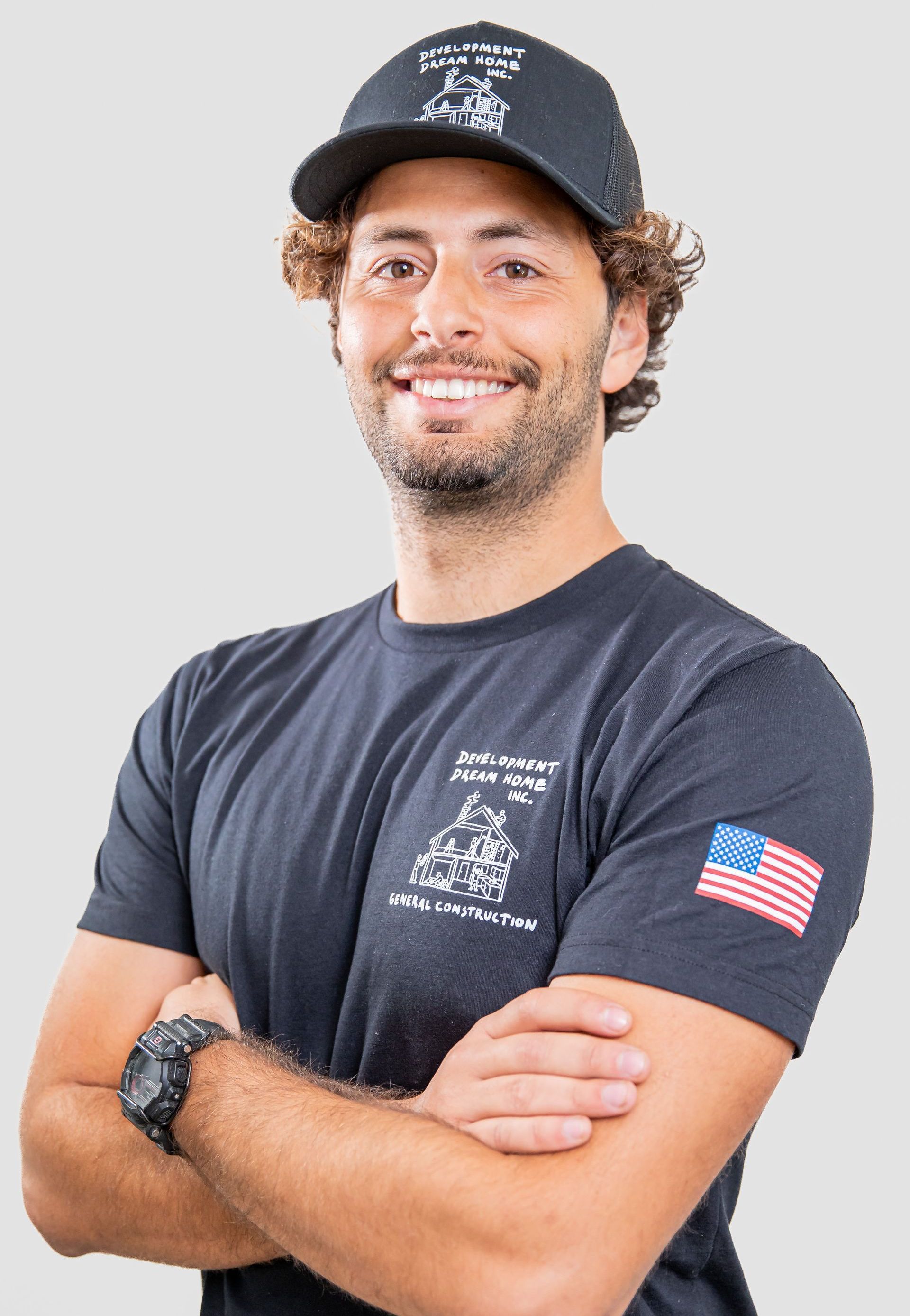 A man wearing a hat and a t-shirt is standing with his arms crossed.
