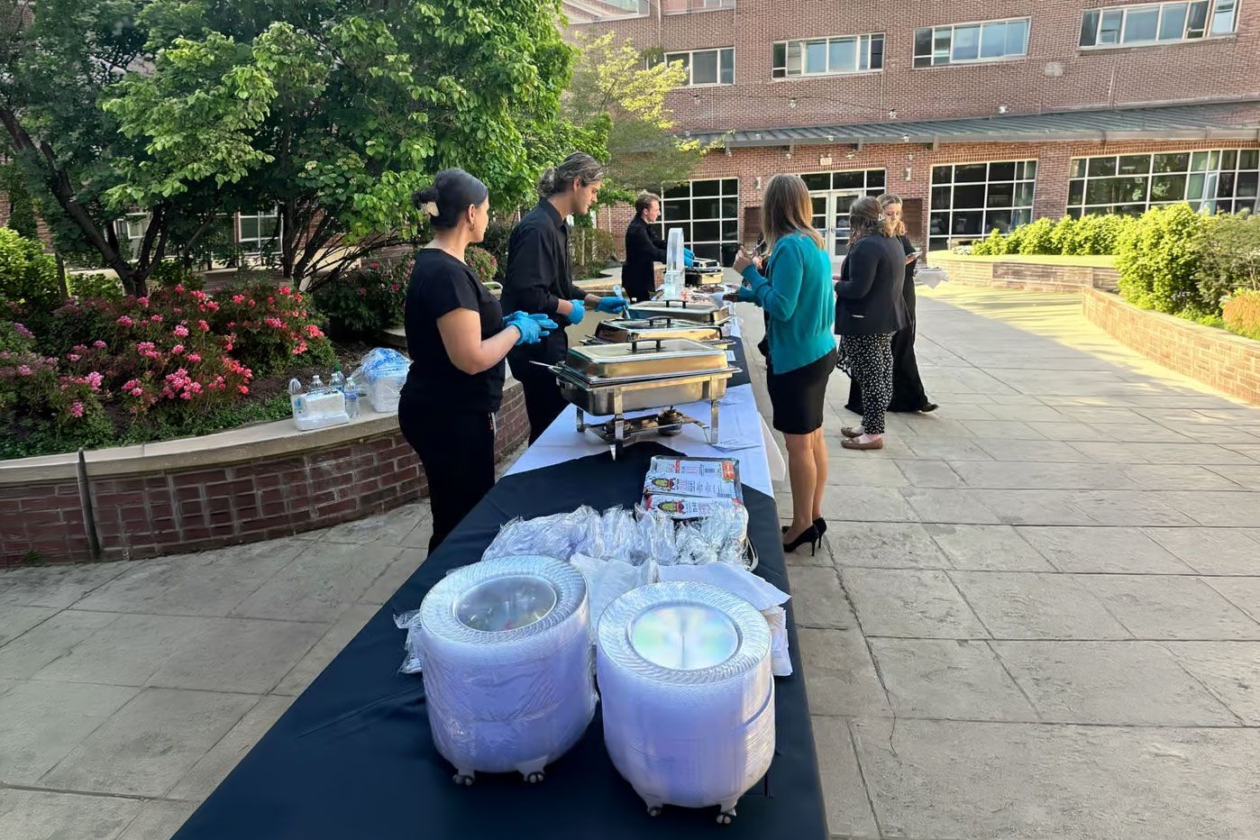 People serving food and drinks at an outdoor campus table with black tablecloths and white beverage tubs.