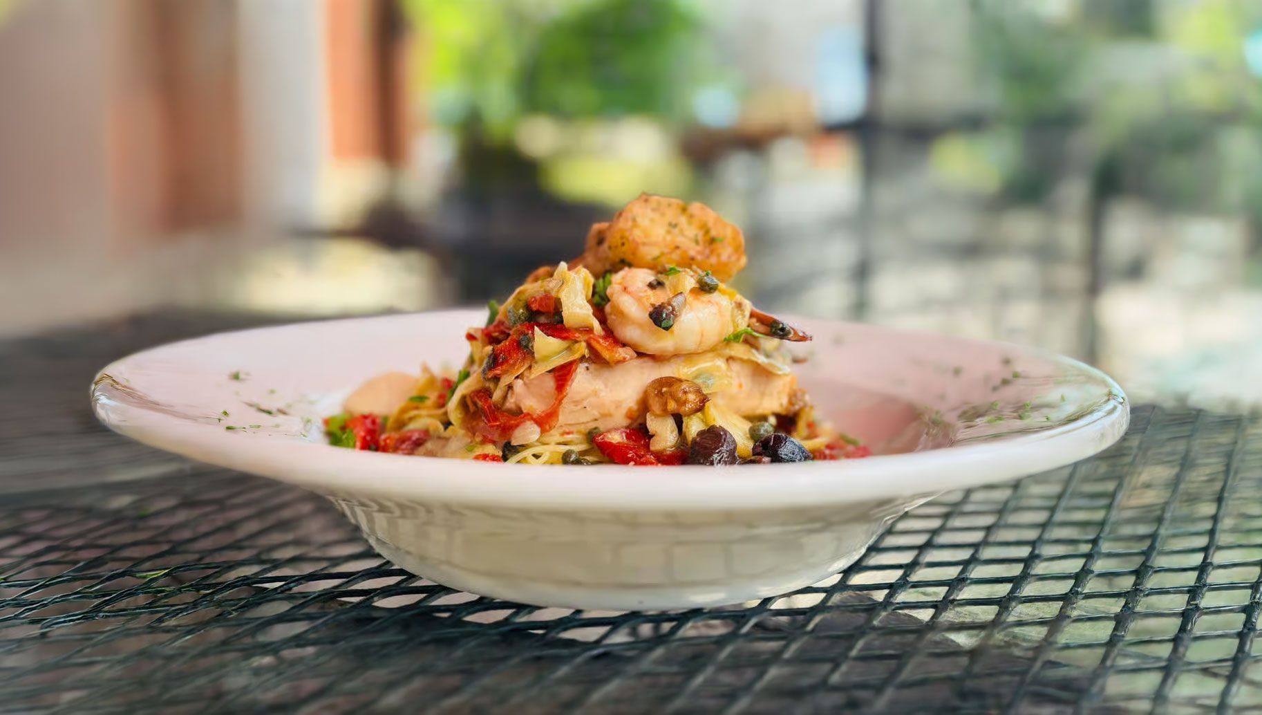 Plated gourmet pasta topped with vegetables and crumbs on a metal patio table, with a blurred outdoor café background