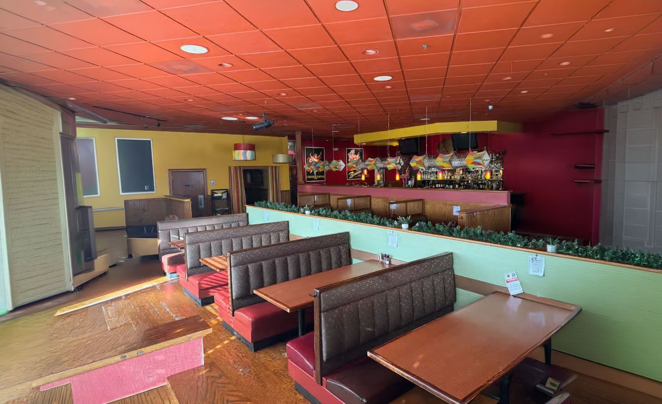 Empty restaurant dining area with booths, tables, and a red ceiling bar counter in the background