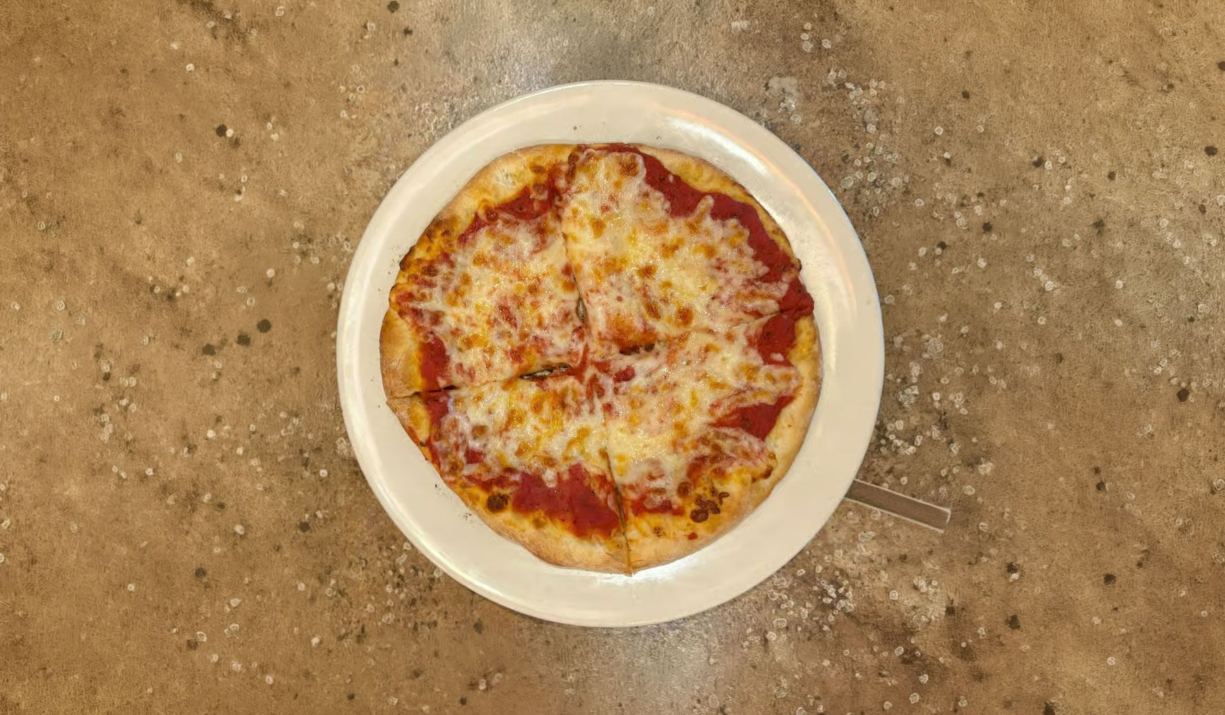 A cheese pizza on a white plate on a tan stone surface.