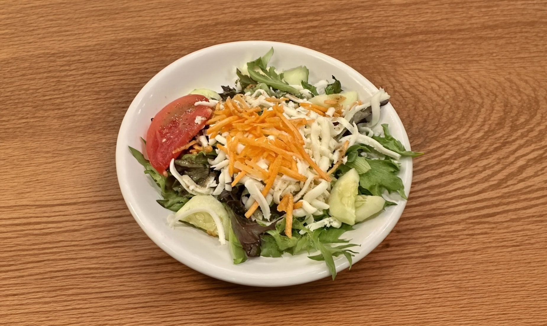 Small salad in a white bowl with lettuce, tomato, avocado, shredded carrots, and cheese on a wooden table