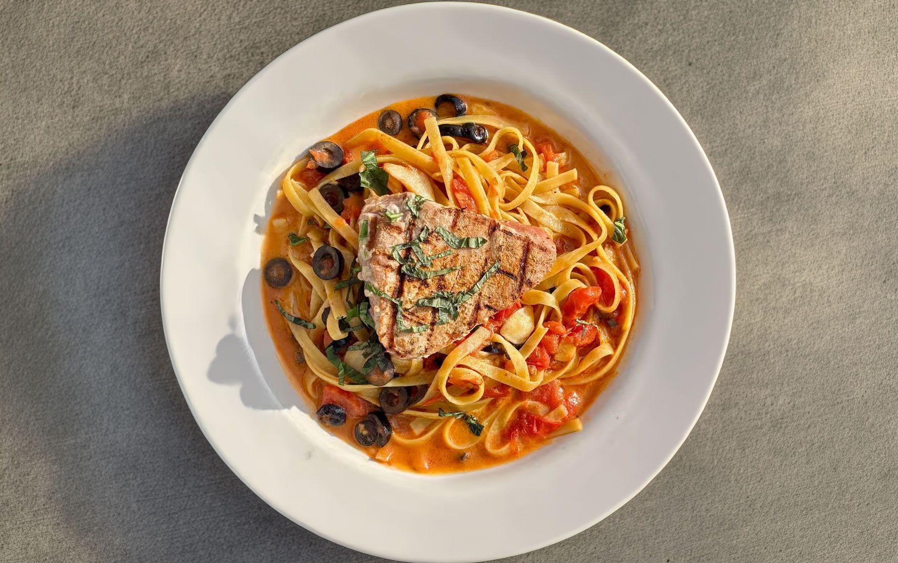 Pasta with tomato sauce, olives, and herbs served in a white bowl on a light surface.