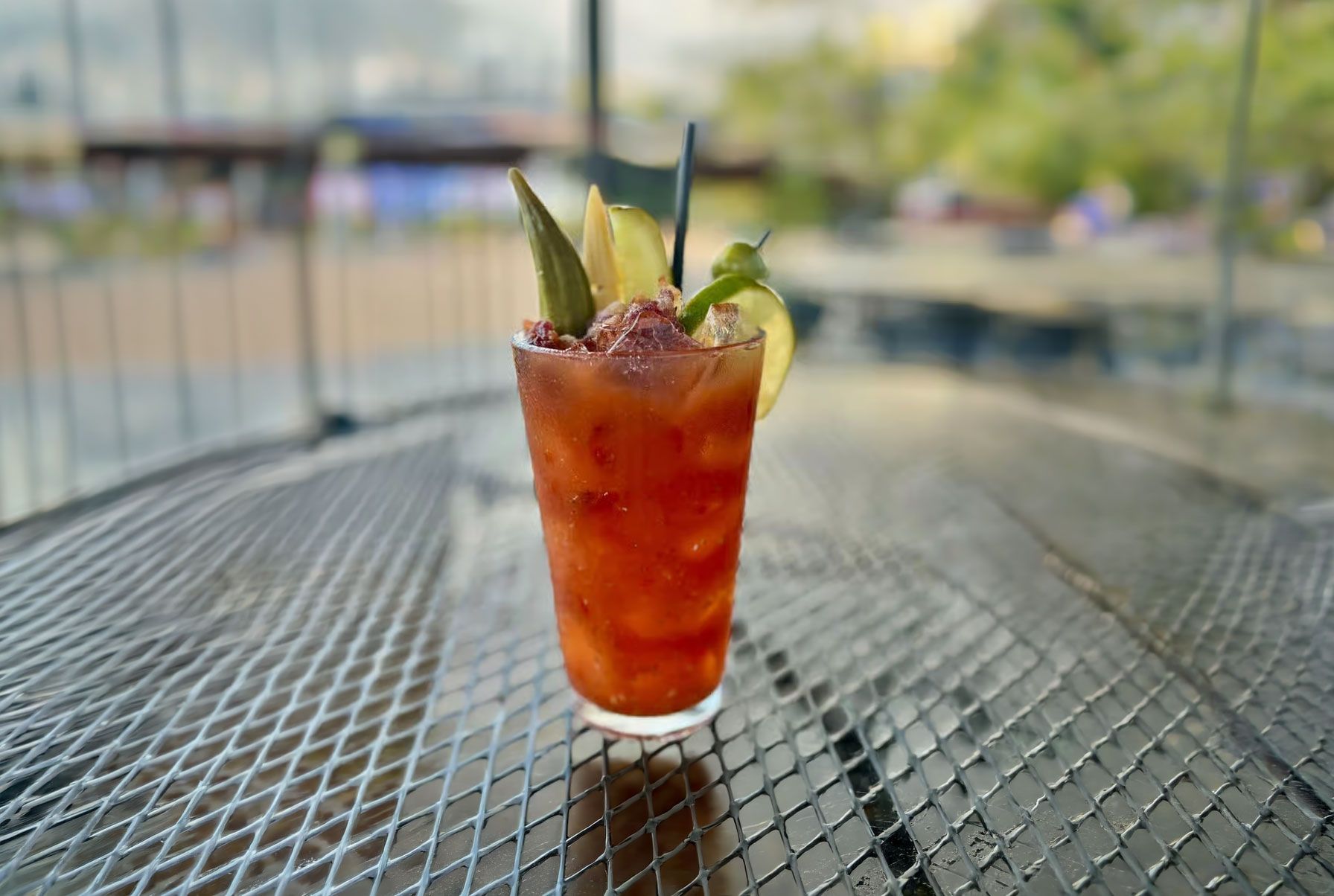 Tall amber cocktail with lime and herbs on a metal outdoor table, blurred waterfront background.
