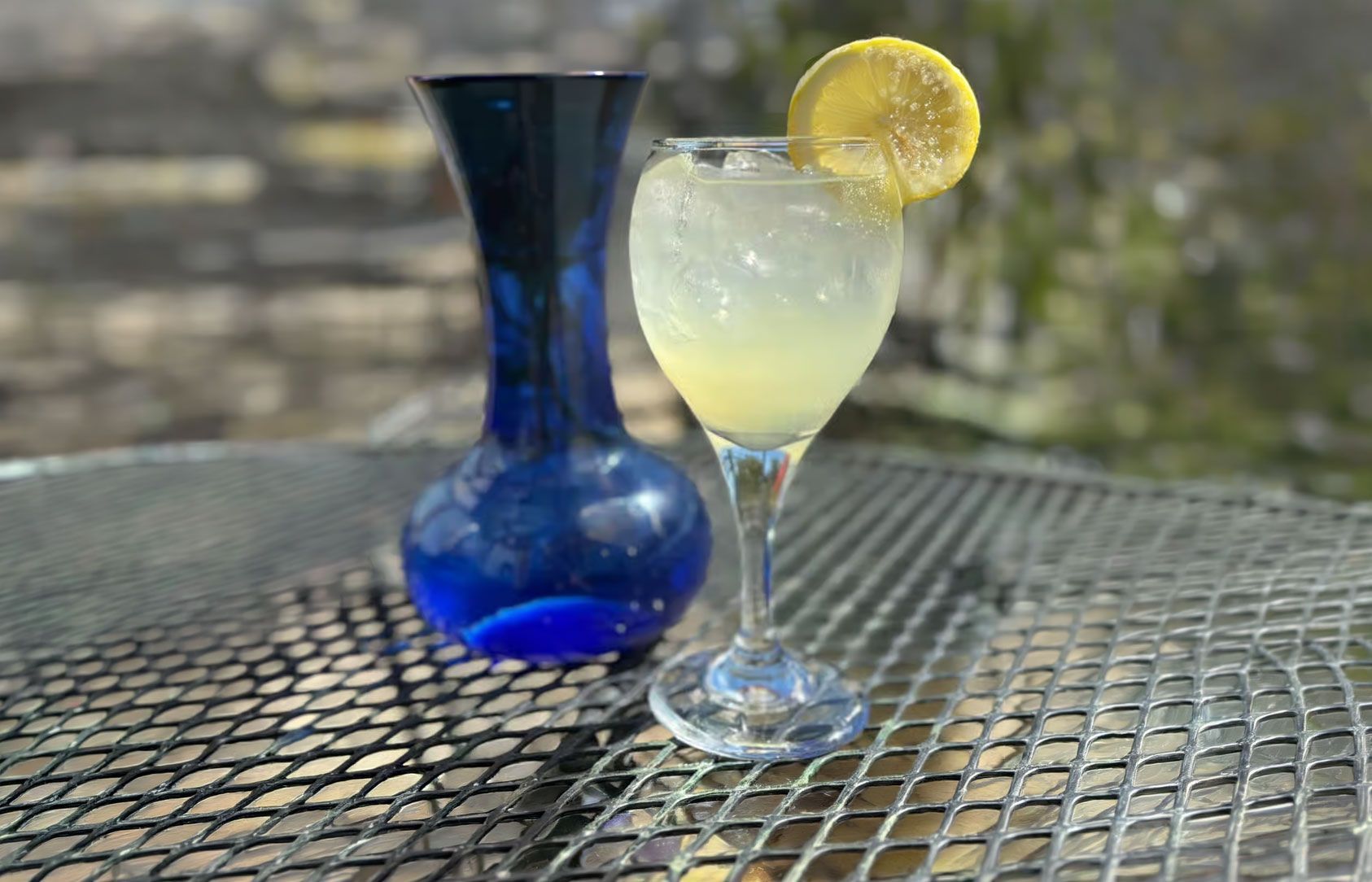 Blue vase and lemon drink in a glass on a metal mesh table outdoors.