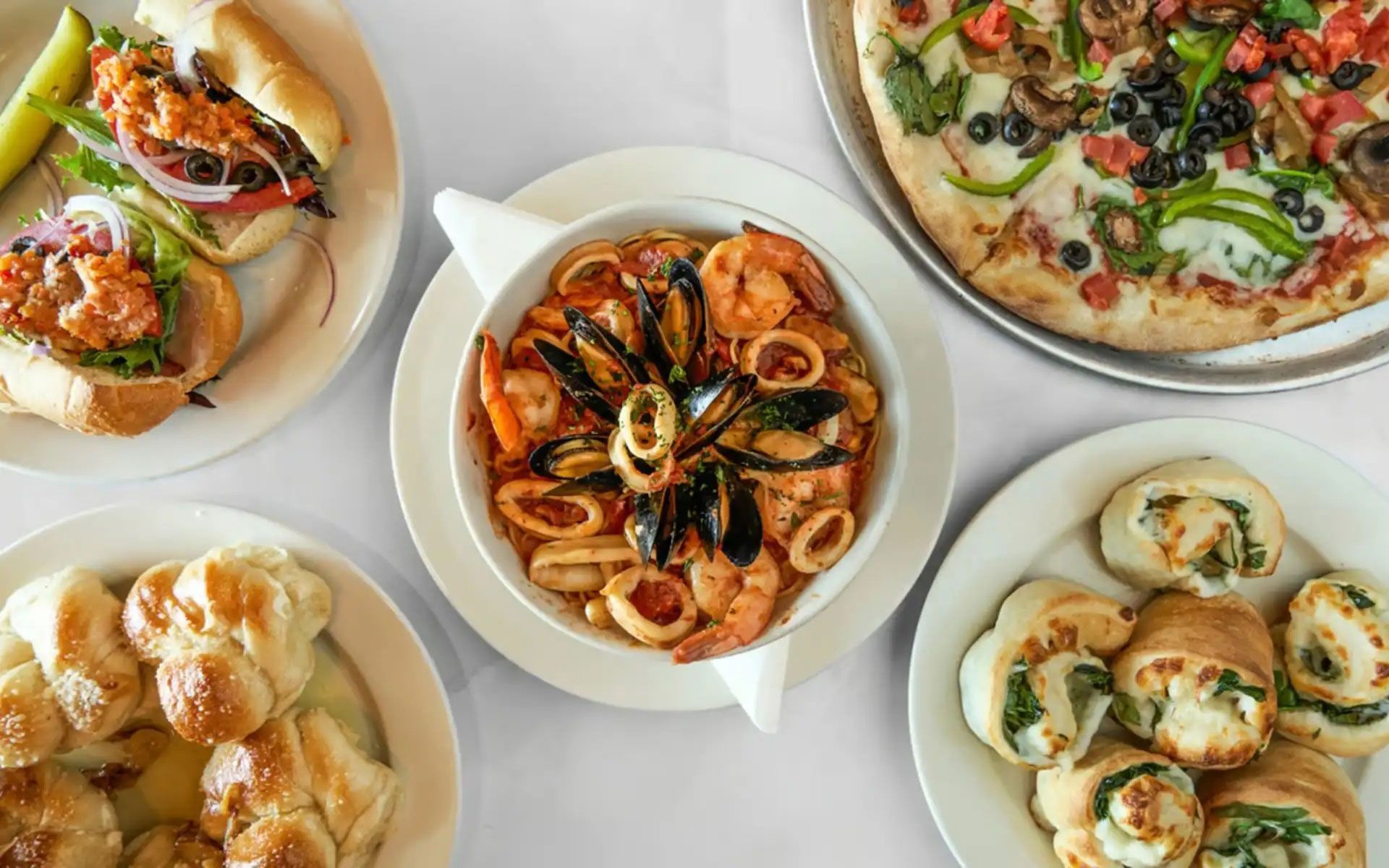 Assorted gourmet dishes on a white tabletop, including pizza, pasta, stuffed rolls, and baked appetizers.