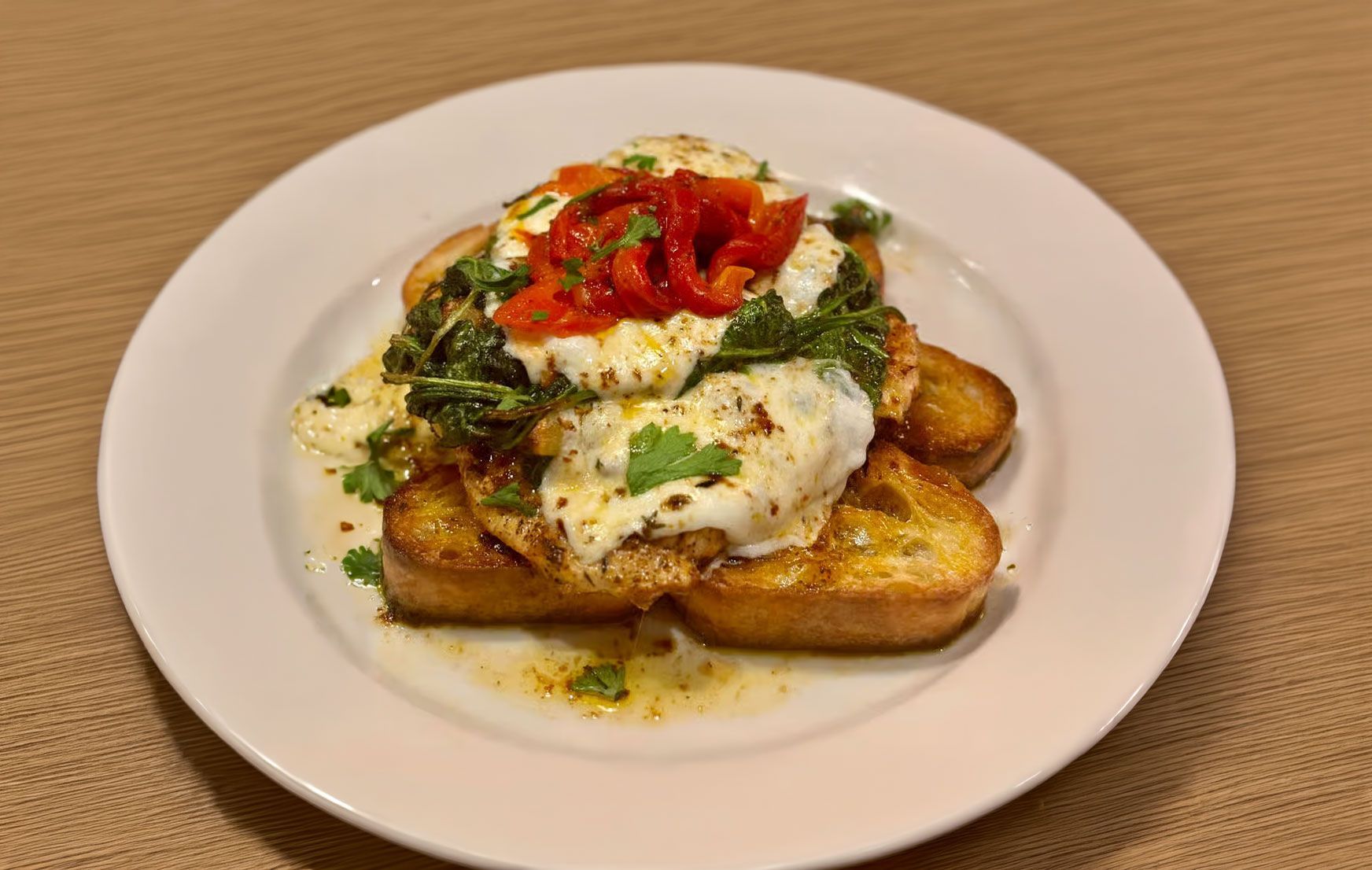 Toast topped with chicken parmesan, greens, and red pepper on a white plate