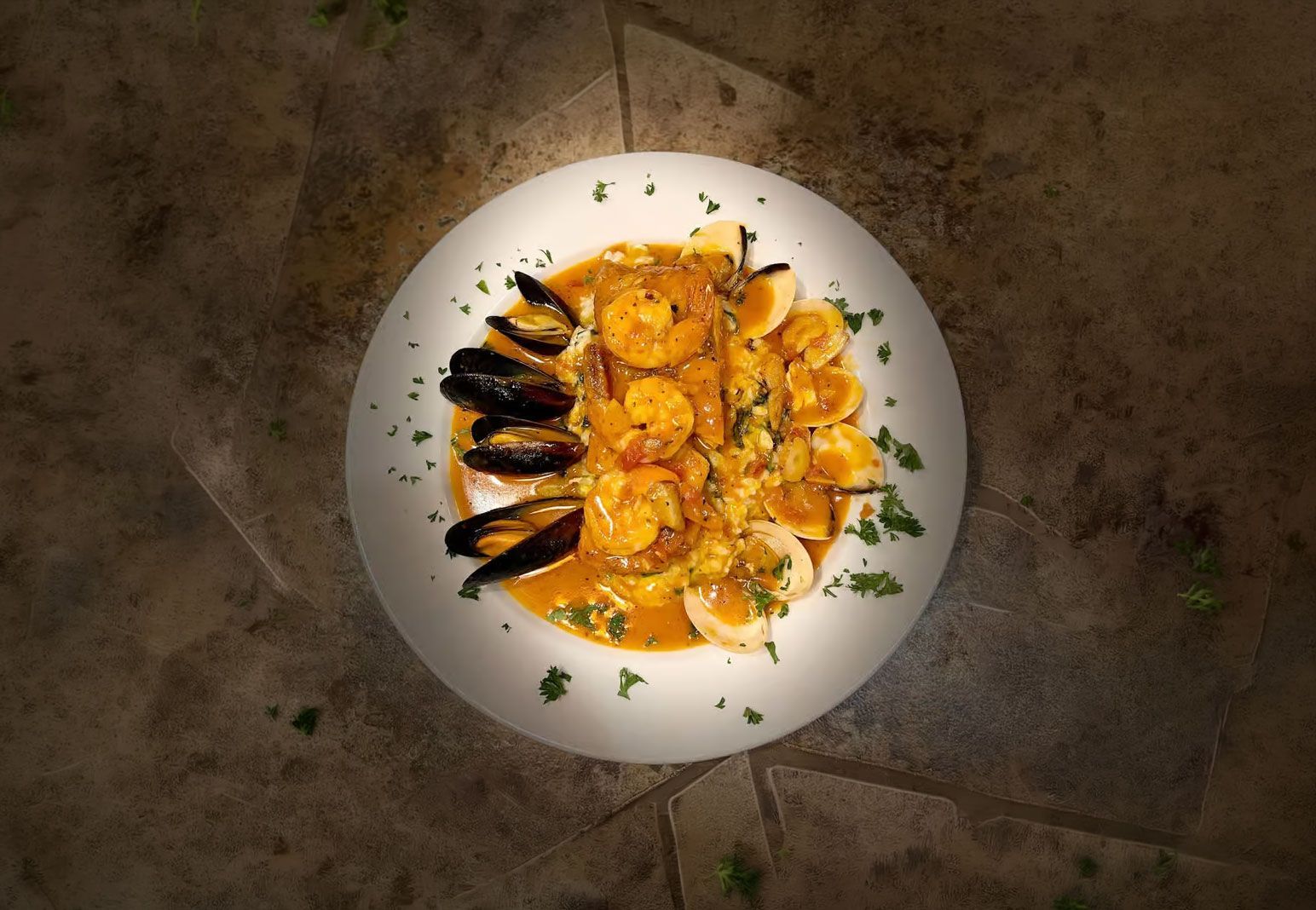 Overhead view of a white plate with shrimp and mussels in orange sauce on a stone table.