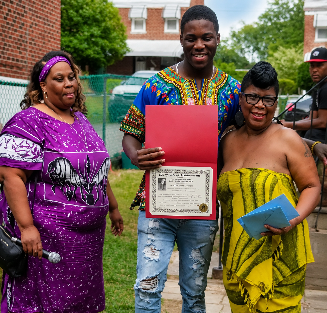 Three people standing outdoors, one holding a certificate in a red folder, smiling in colorful clothing.