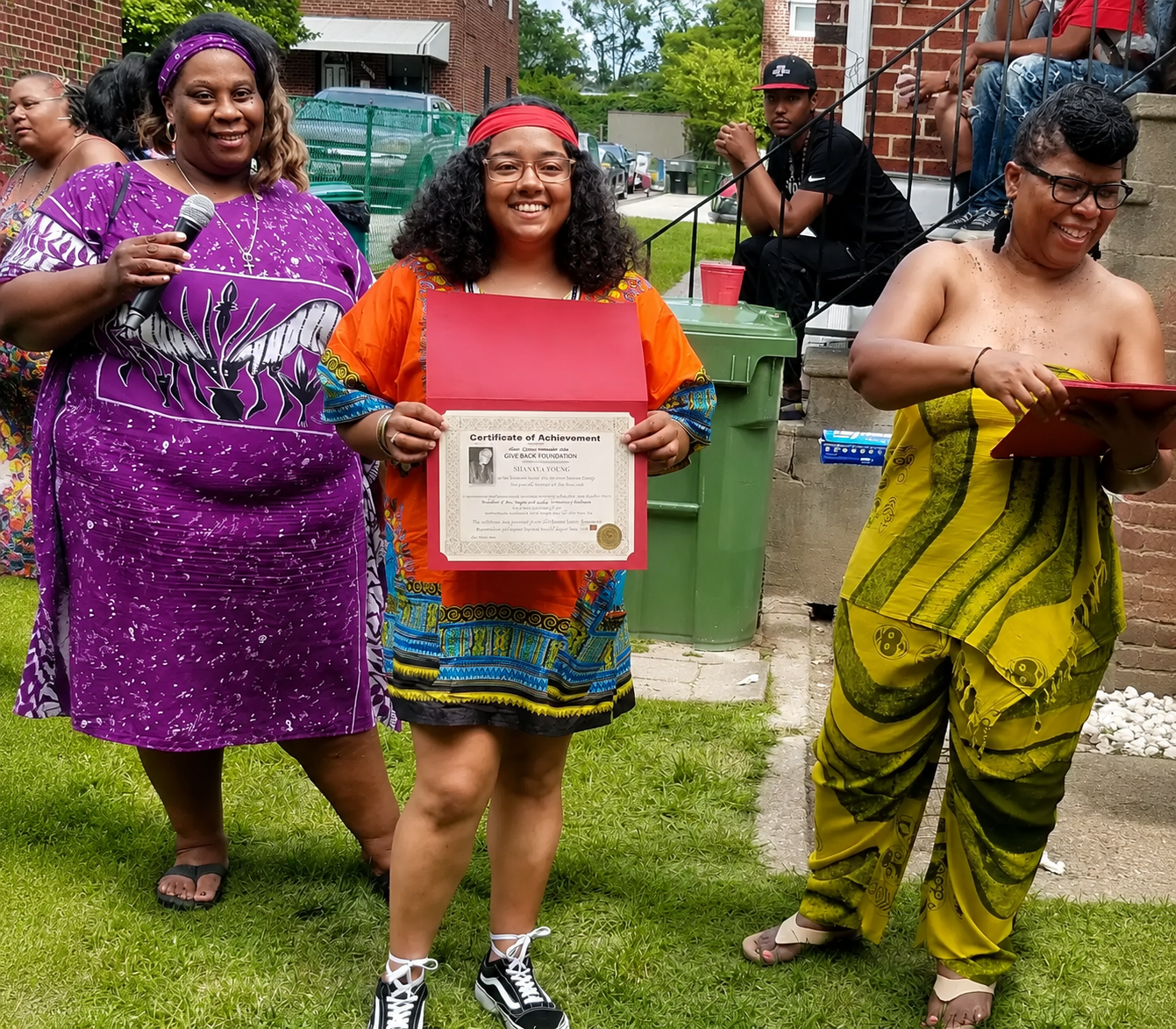 Three people in colorful dresses smile outdoors at a celebration, one holding a framed certificate.