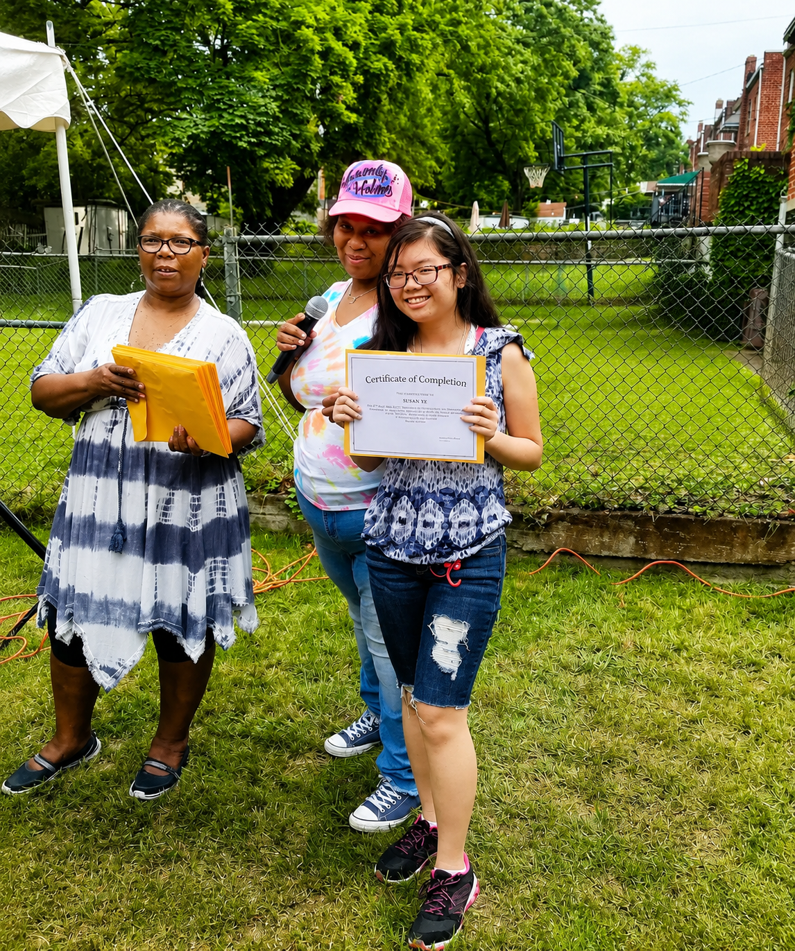 Three people posing outdoors on grass, two holding papers, near a chain-link fence and trees.