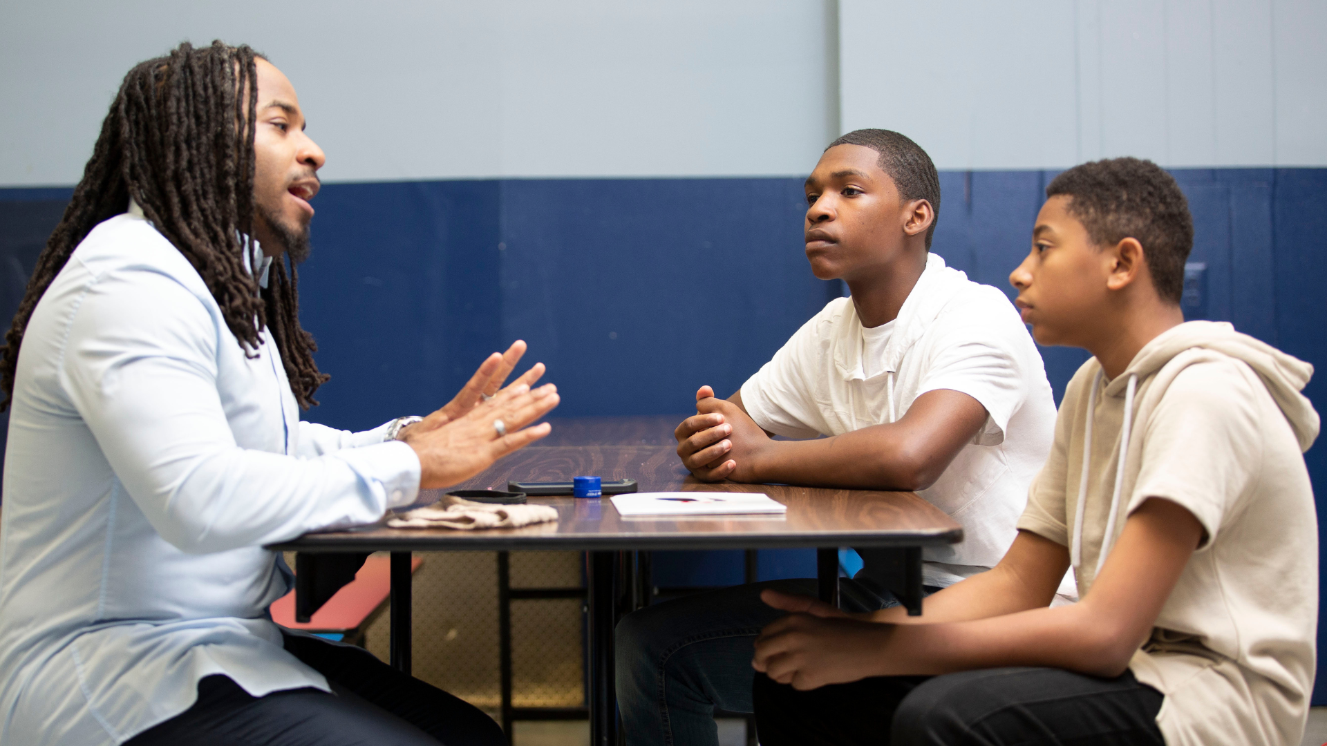 Teacher and two students talking at a table in a classroom, with notebooks and a blue wall behind them.