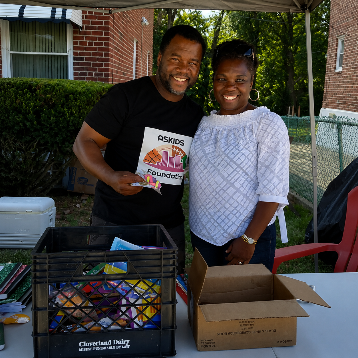 Two smiling people at an outdoor table with boxes and a crate of supplies under a canopy.
