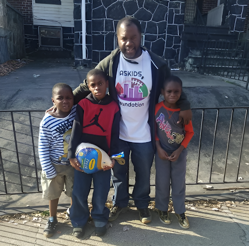 Four people standing together outdoors in front of a house gate, holding a soccer ball.