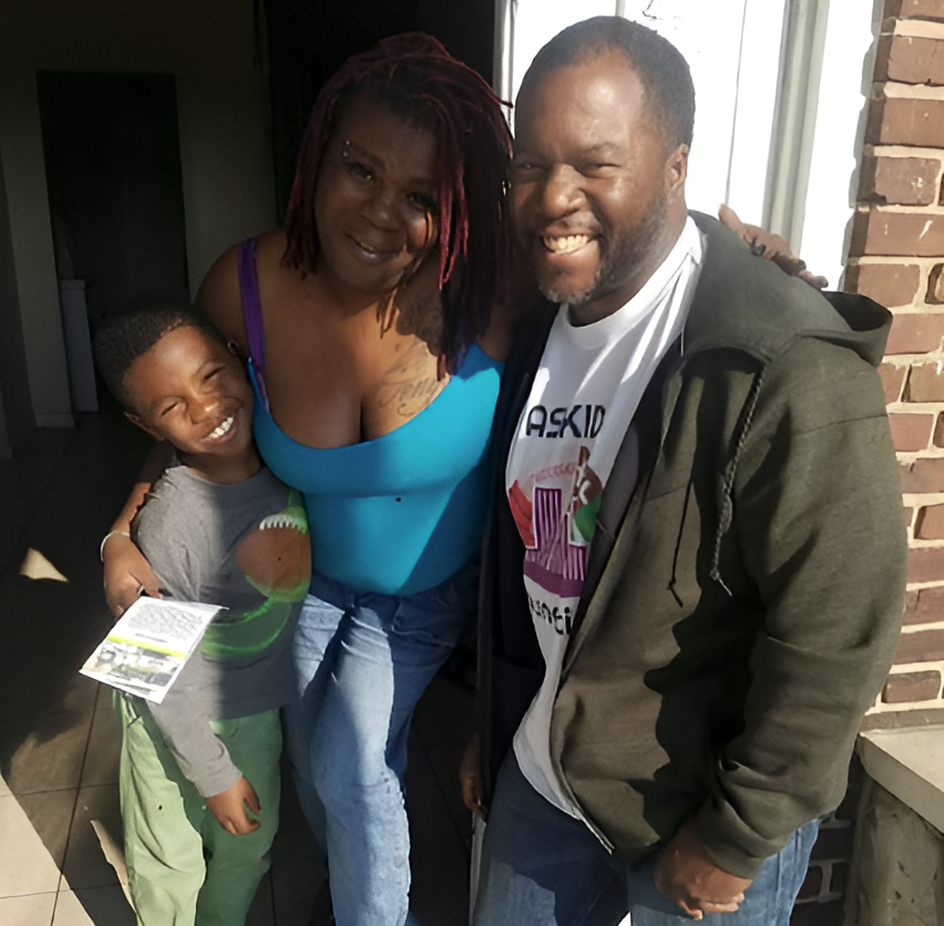 Three people smiling on a porch in bright sunlight, one holding a paper, another in a blue top.