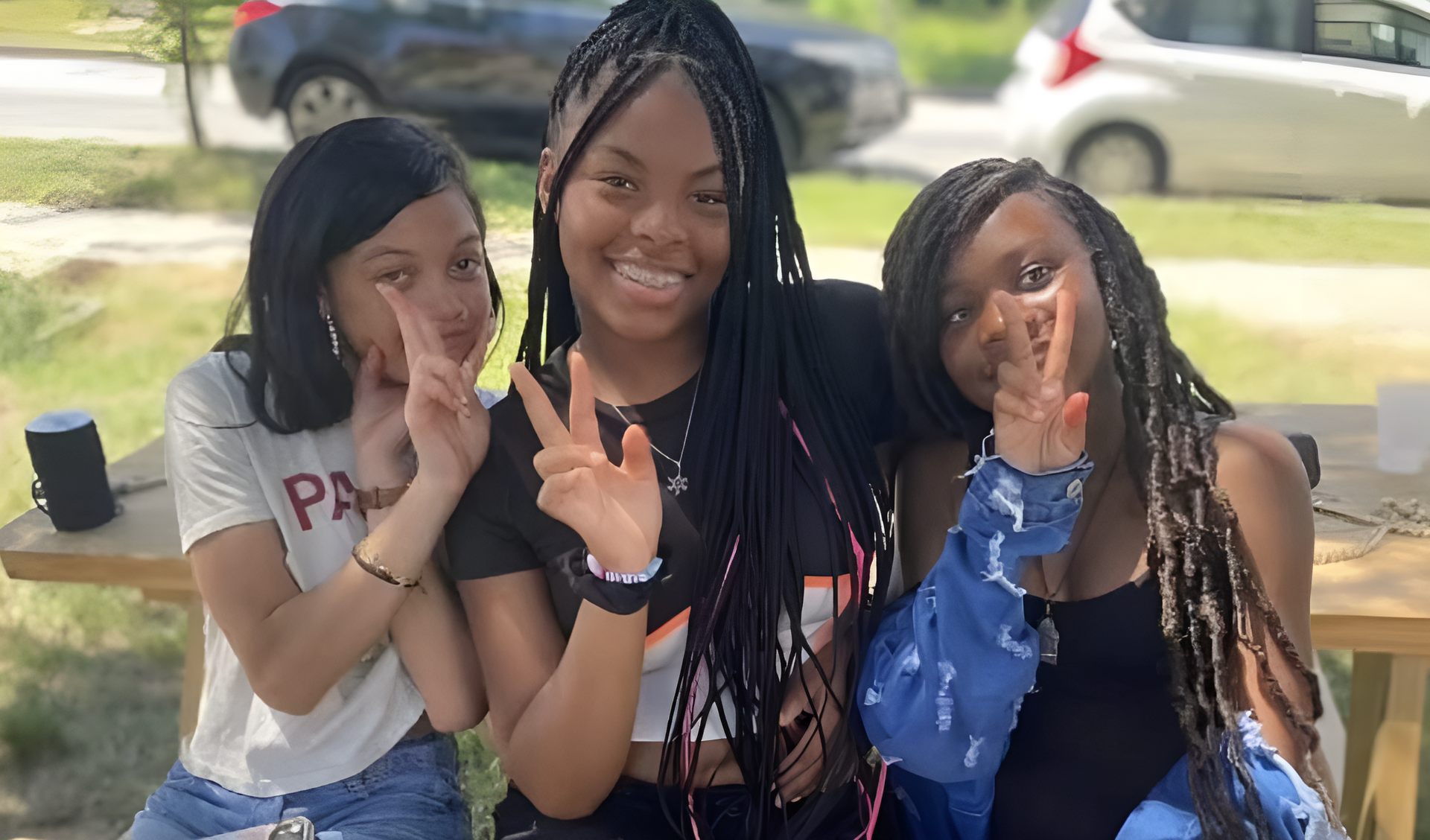 Three friends sitting outdoors, smiling and posing with peace signs under a shaded area.