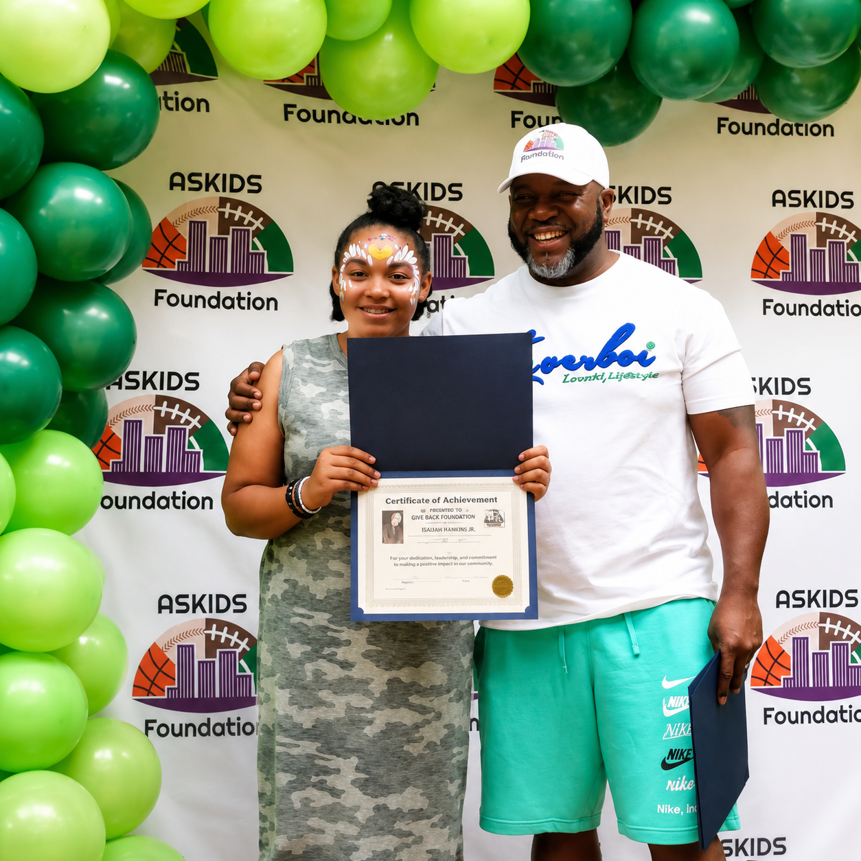 Two people pose with a certificate in front of a green balloon wall and ASKIDS Foundation backdrop.