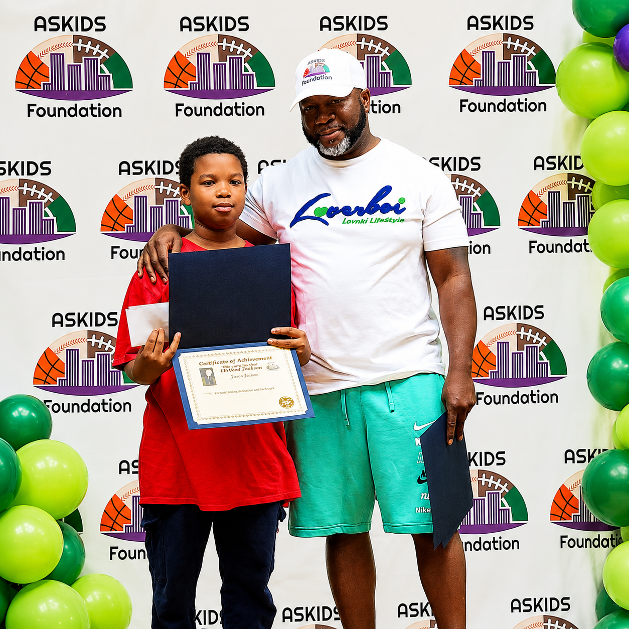 Boy holding a certificate beside a man at an ASKIDS Foundation event with balloons and a logo backdrop