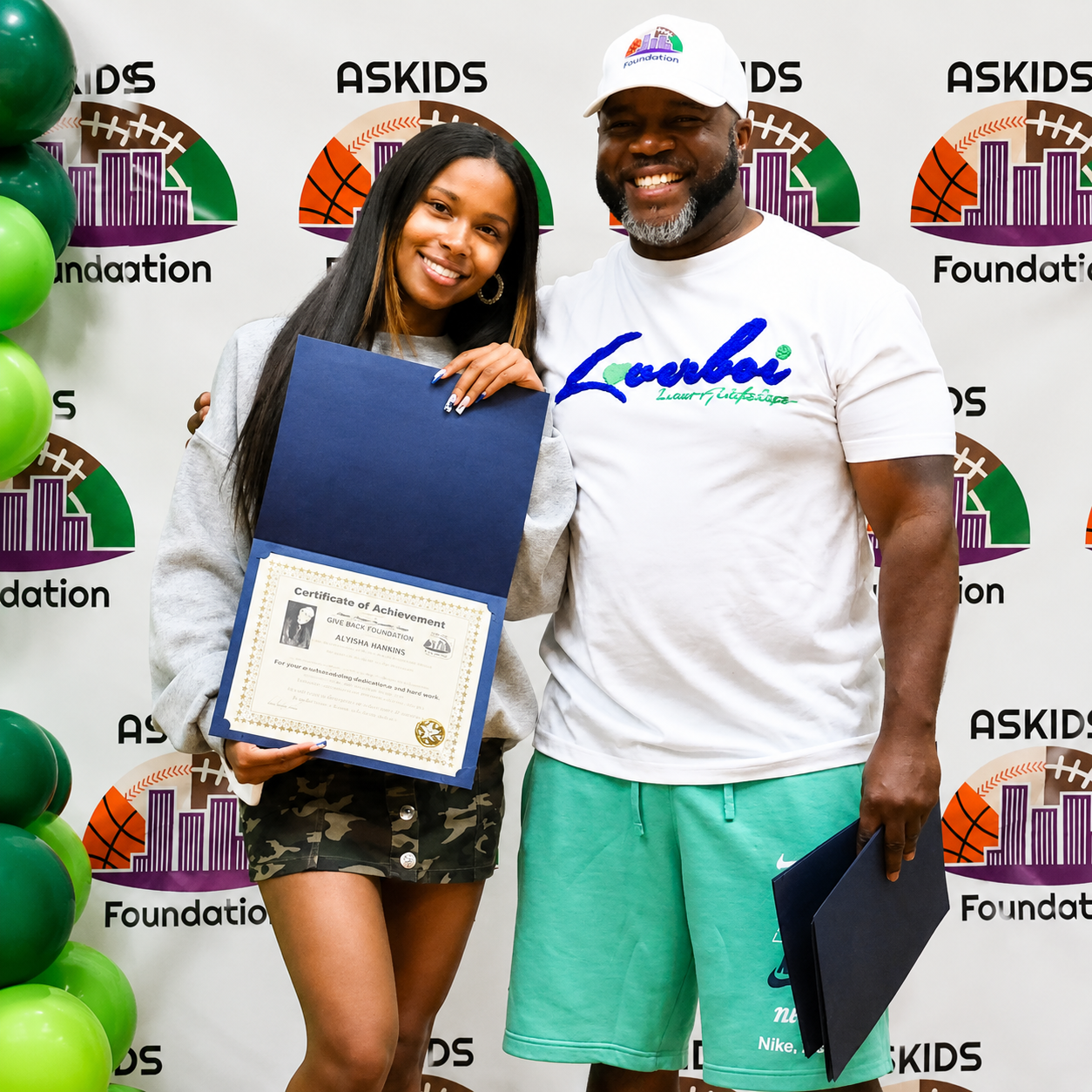 Two people holding certificates in front of an ASKIDS Foundation backdrop, smiling at an event.
