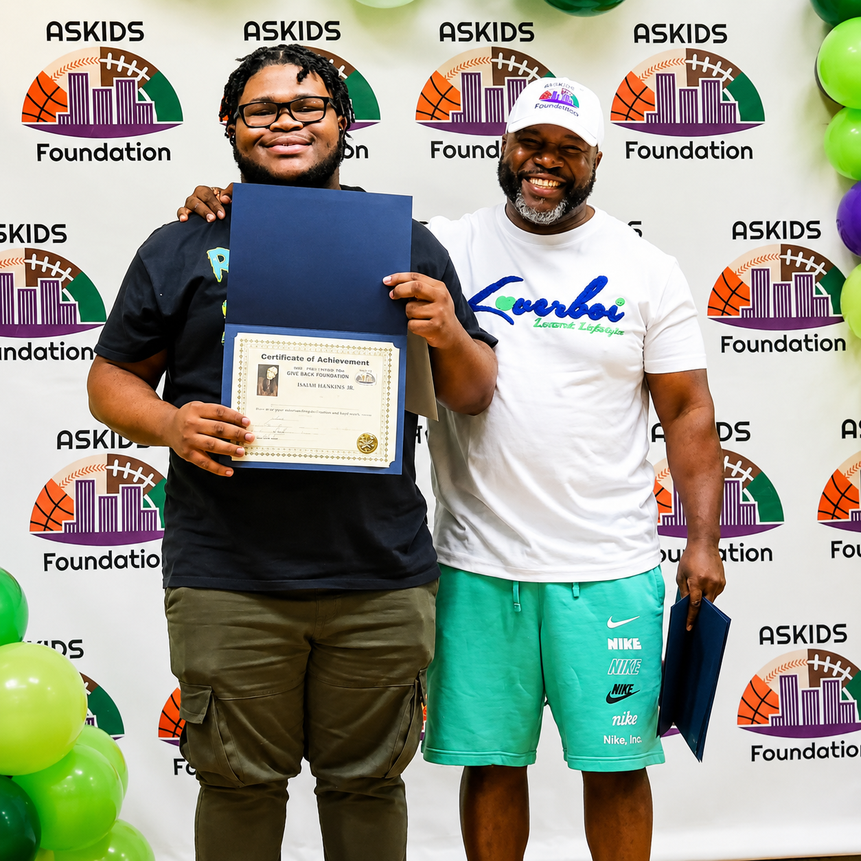 Two men pose at an ASKIDS Foundation event, one holding a certificate, with balloons in the corner.
