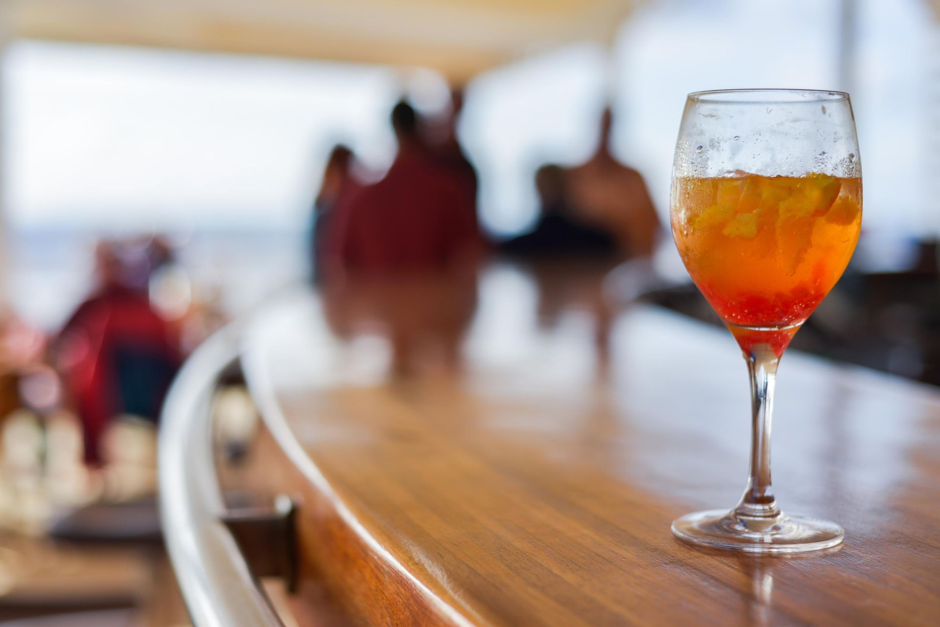 Orange cocktail in a stemmed glass on a wooden bar with blurred figures in the background.