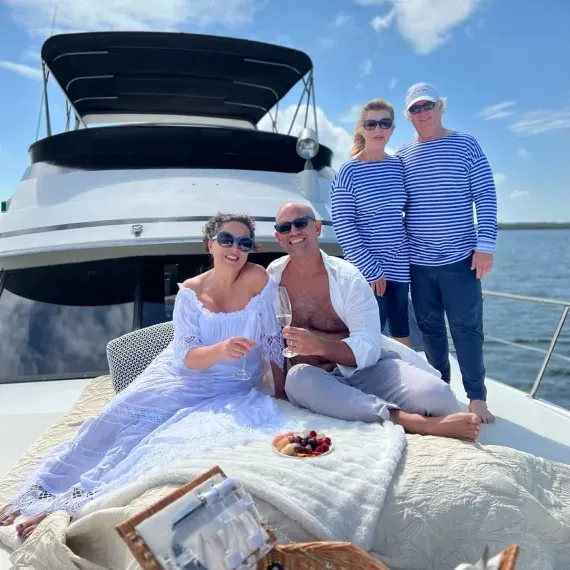 People on a boat enjoying a picnic. Two in blue and white striped shirts, others relaxed on a blanket, smiling. Blue sky.