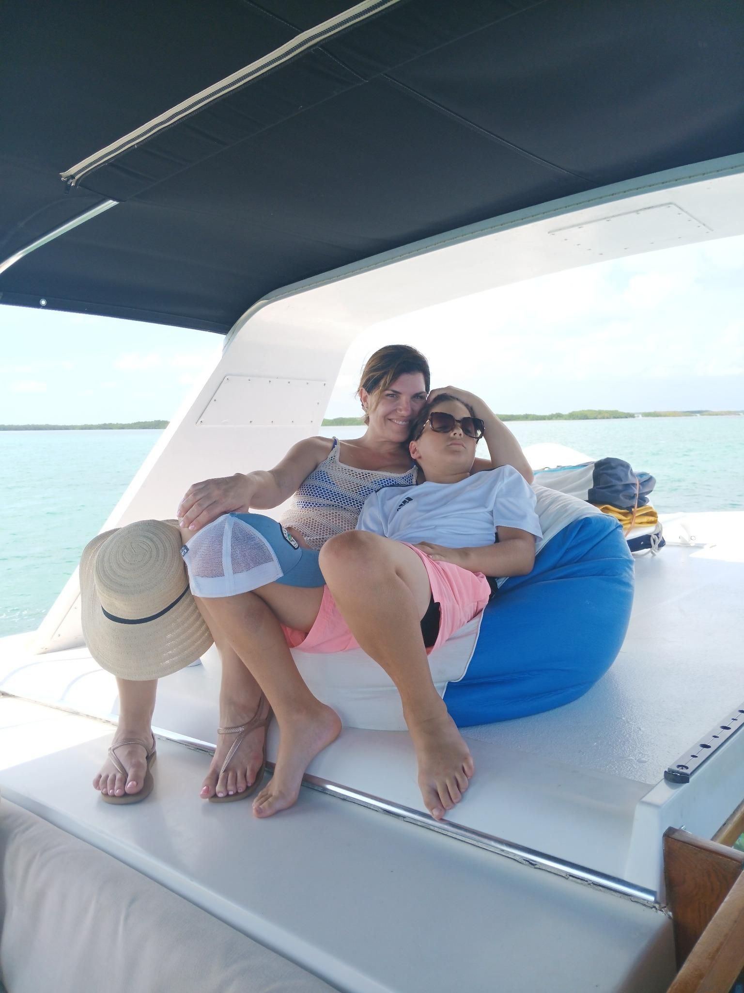 Two women relaxing on a boat; one has arm around the other, both smiling. Blue sky and water in the background.