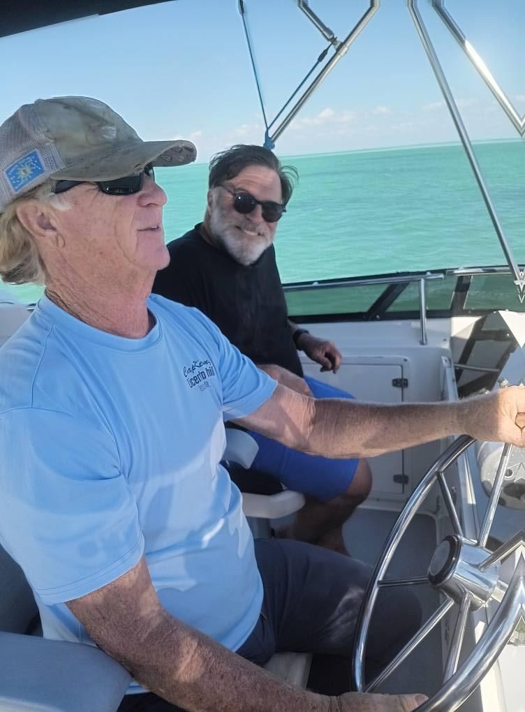 Man in blue shirt steering boat, another man smiling in background.