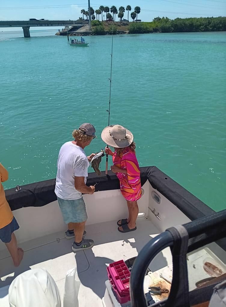People fishing from a boat on blue water, holding a fish. A bridge is in the background.