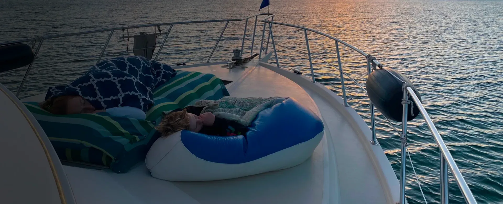 People sleeping on a boat's deck, blankets and beanbag. Ocean in the background. Sunset colors.
