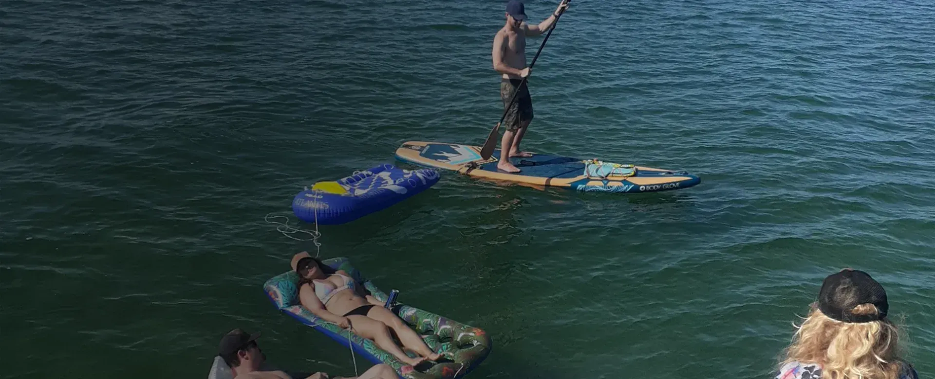 Person paddleboarding on water; others floating on rafts. Sunny day.