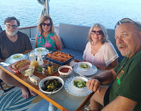 Four people dine on a boat. Food is on the table, ocean in background.