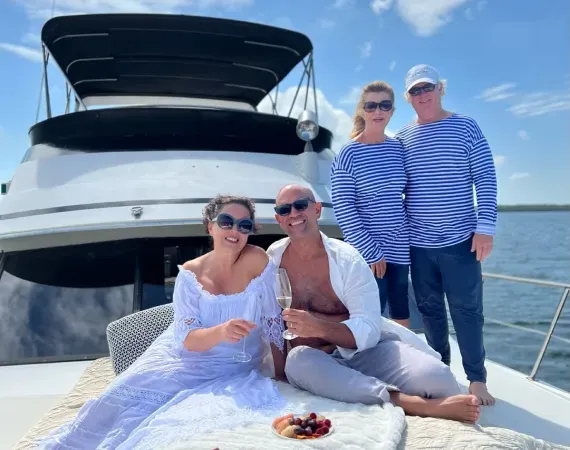 Four people on a yacht. Two sit, holding glasses; others stand, wearing blue-striped tops, under a sunny sky.