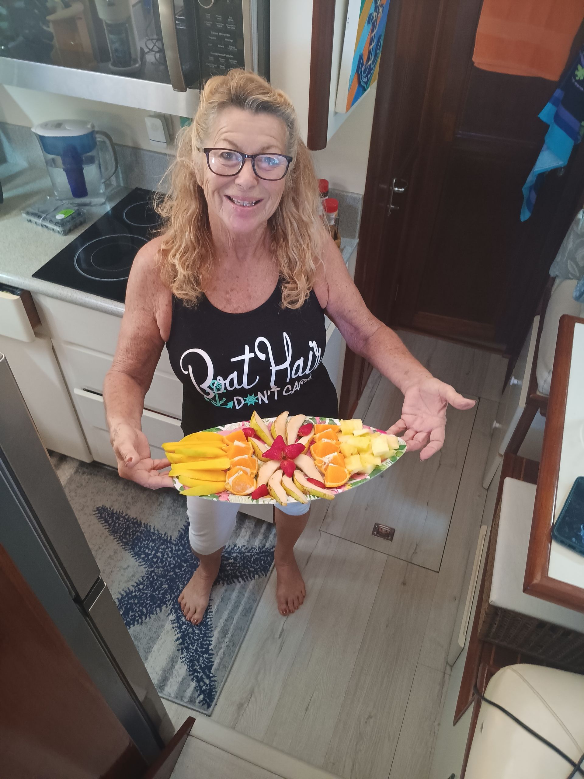 Woman holding a platter of sliced fruit, smiling inside a boat's galley.